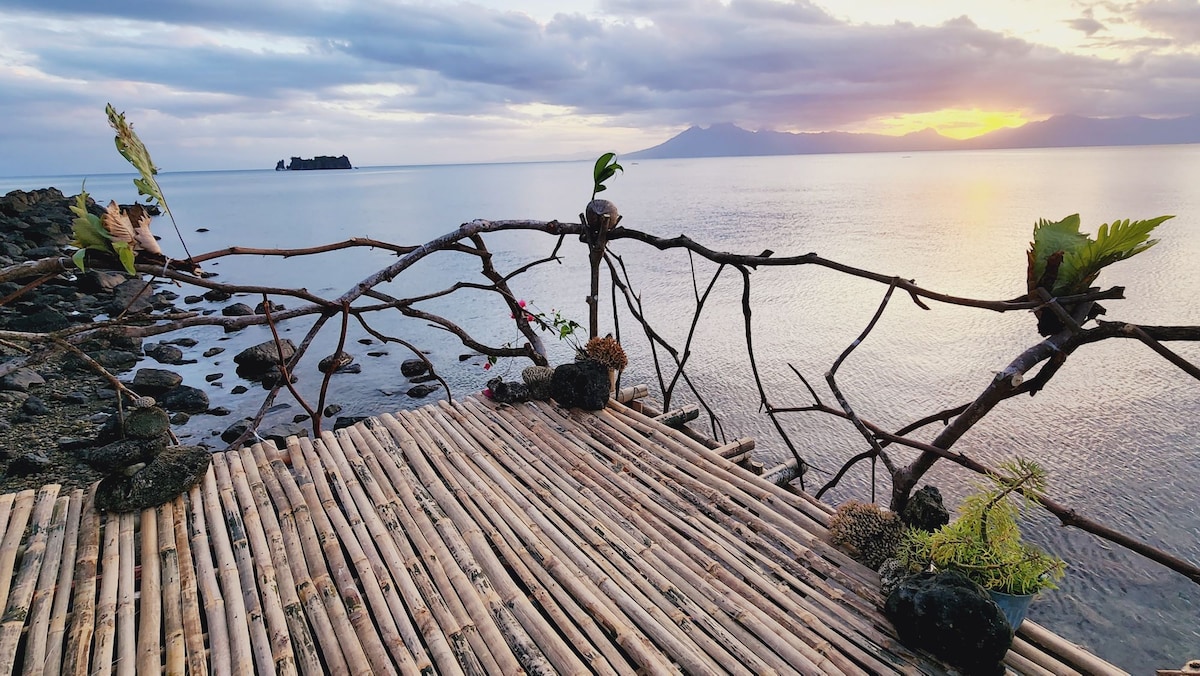 A wooden deck made of bamboo extends over calm waters, with natural rock formations visible along the shoreline. Decorative plants are placed on the deck's edge, complemented by a soft sunset glow reflecting on the water's surface.