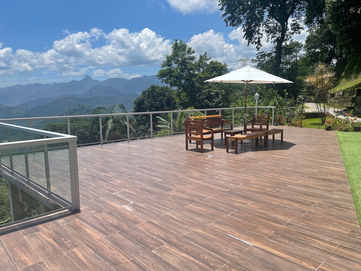 A spacious outdoor deck offers a panoramic view of the surrounding mountains. Several wooden chairs and a coffee table are arranged under a large umbrella, providing a comfortable seating area. Lush greenery and vibrant flowers are visible in the background.