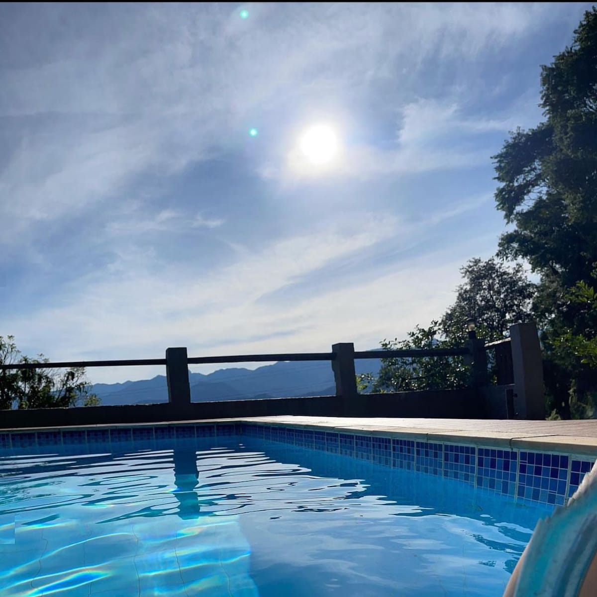 The image captures a clear swimming pool reflecting sunlight, with a view of distant mountains under a bright sky. Lush greenery frames the scene, and a railing provides a boundary between the pool area and the surrounding landscape.