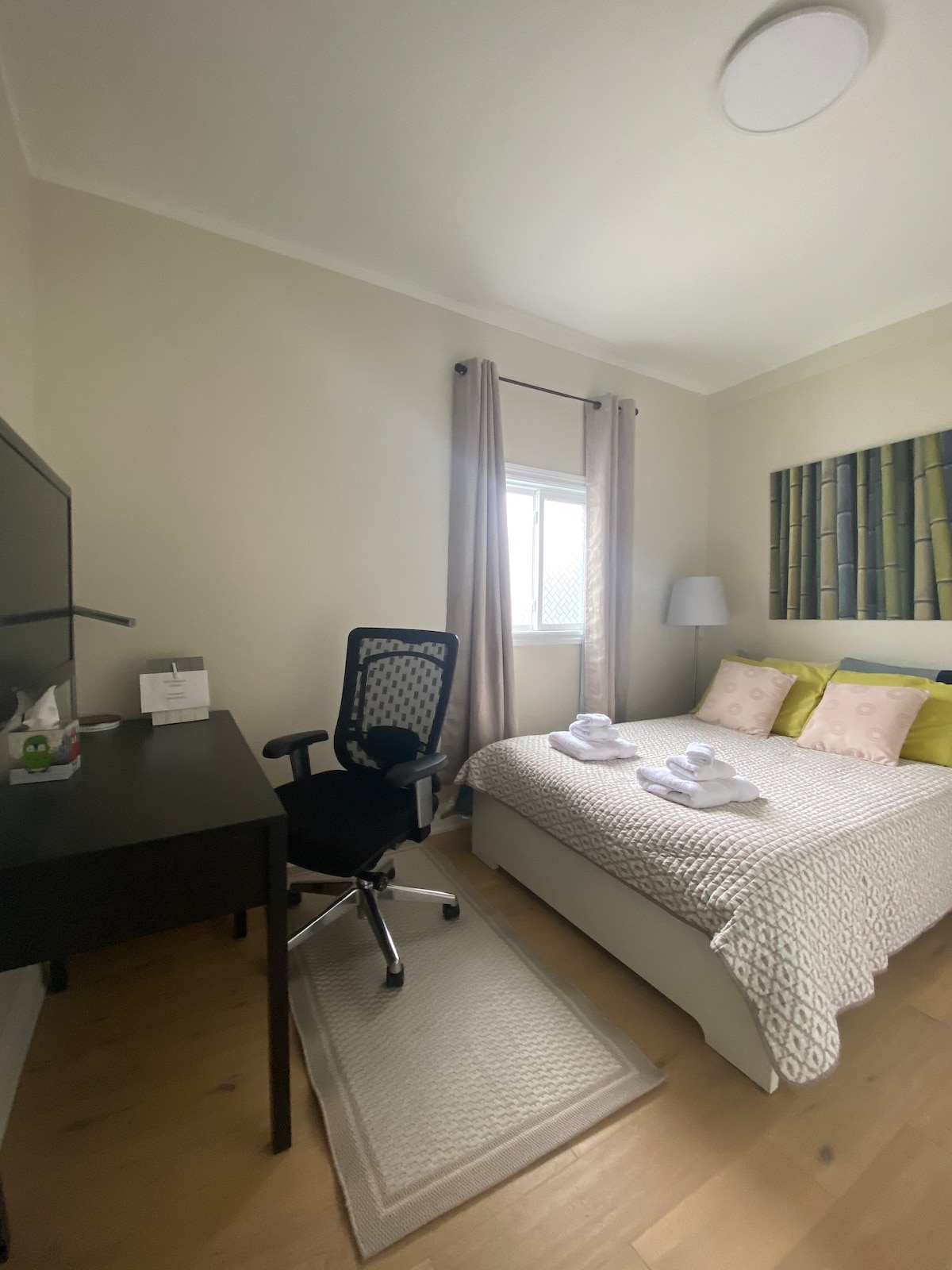 The bedroom features a double bed with soft linens and towels neatly arranged. A contemporary desk with an ergonomic chair is positioned nearby. Natural light filters through the window, while a piece of artwork depicting bamboo adds a serene touch.