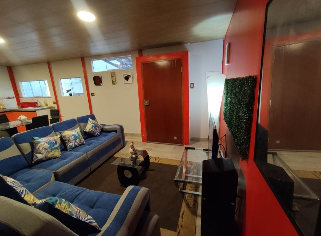 A spacious living area features a comfortable blue sofa adorned with tropical-patterned cushions. A glass coffee table rests on a neutral rug in front of a wall-mounted television. Bright walls provide a vibrant contrast to the overall decor.