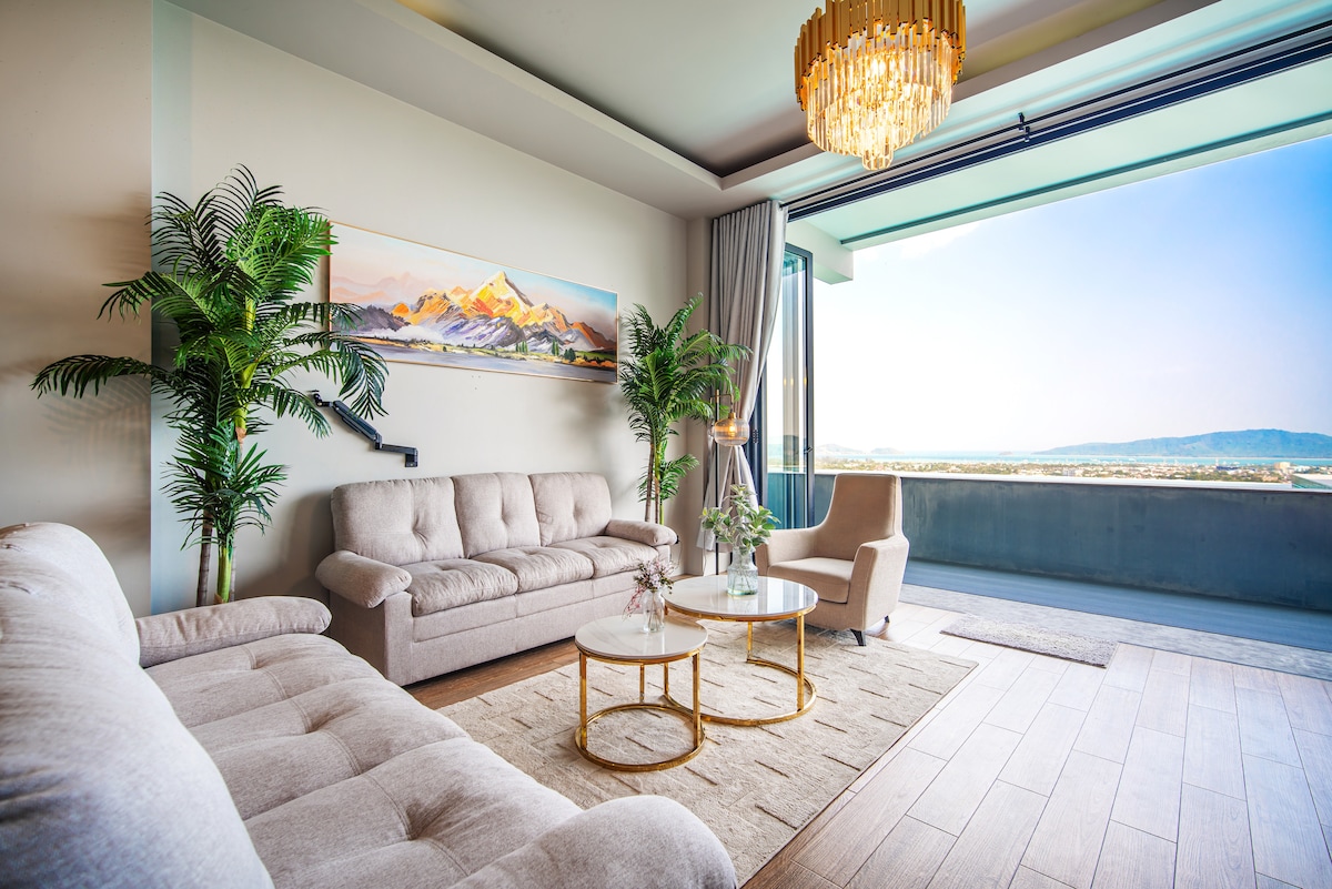 A bright living area features a large sliding glass door, leading to a scenic view of the mountains and sea. Comfortable beige sofas are arranged around a round coffee table, complemented by decorative plants and soft lighting from a chandelier.
