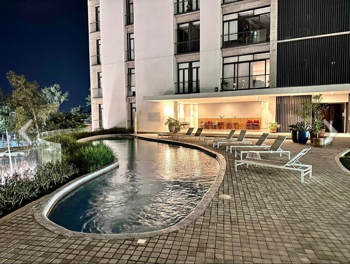 A serene pool area is set against the backdrop of a modern building, featuring gently curved edges and a tranquil water surface. Lounge chairs are arranged along the poolside, with lush greenery enhancing the outdoor space. Soft illumination creates a calming evening ambiance.