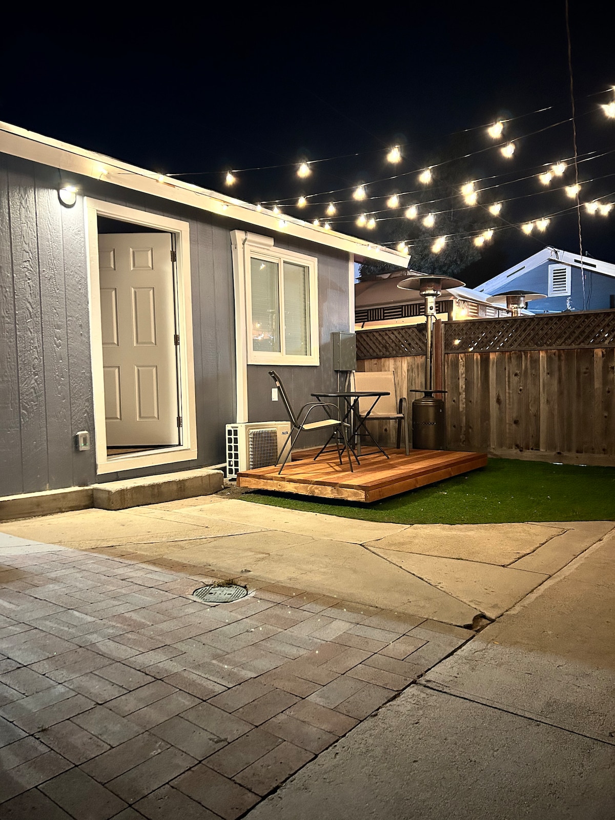 A modern studio exterior is illuminated by string lights, creating a welcoming ambiance. The private patio features a small table and chairs on a wooden deck overlooking a turf grass area. A door leads into the studio, with a nearby outdoor heater for comfort.
