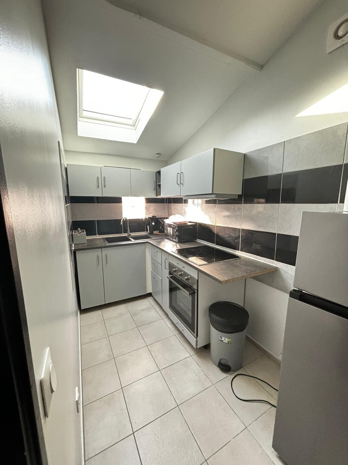 A functional kitchen is equipped with modern cabinetry and a countertop. A skylight overhead allows natural light to illuminate the space. Kitchen appliances, including a stove and a refrigerator, are neatly arranged. The tiled floor complements the overall clean design, enhancing usability.