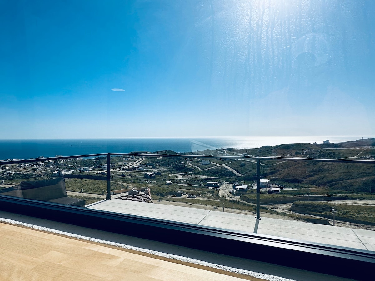 A sweeping view of the ocean stretches beyond the expansive glass railing. The coastline and rolling hills are visible under a clear blue sky, highlighting the natural beauty of the area and creating an inviting sense of tranquility.