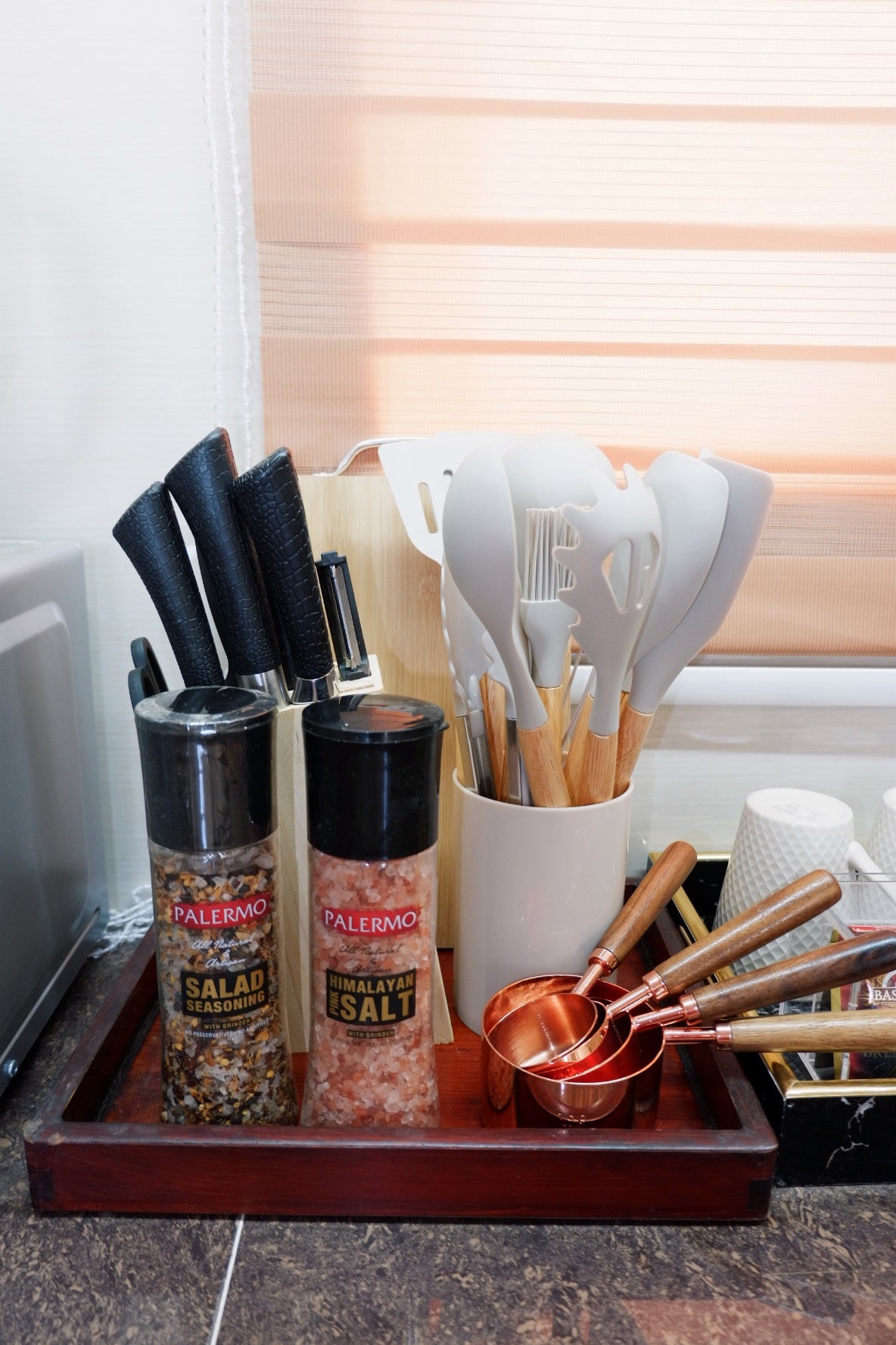 A well-organized kitchen counter displays essential cooking tools. Knife sets are stored in a holder alongside salt and salad seasoning containers. Various utensils, including spatulas and measuring cups, are neatly arranged in a container, contributing to a functional cooking space.