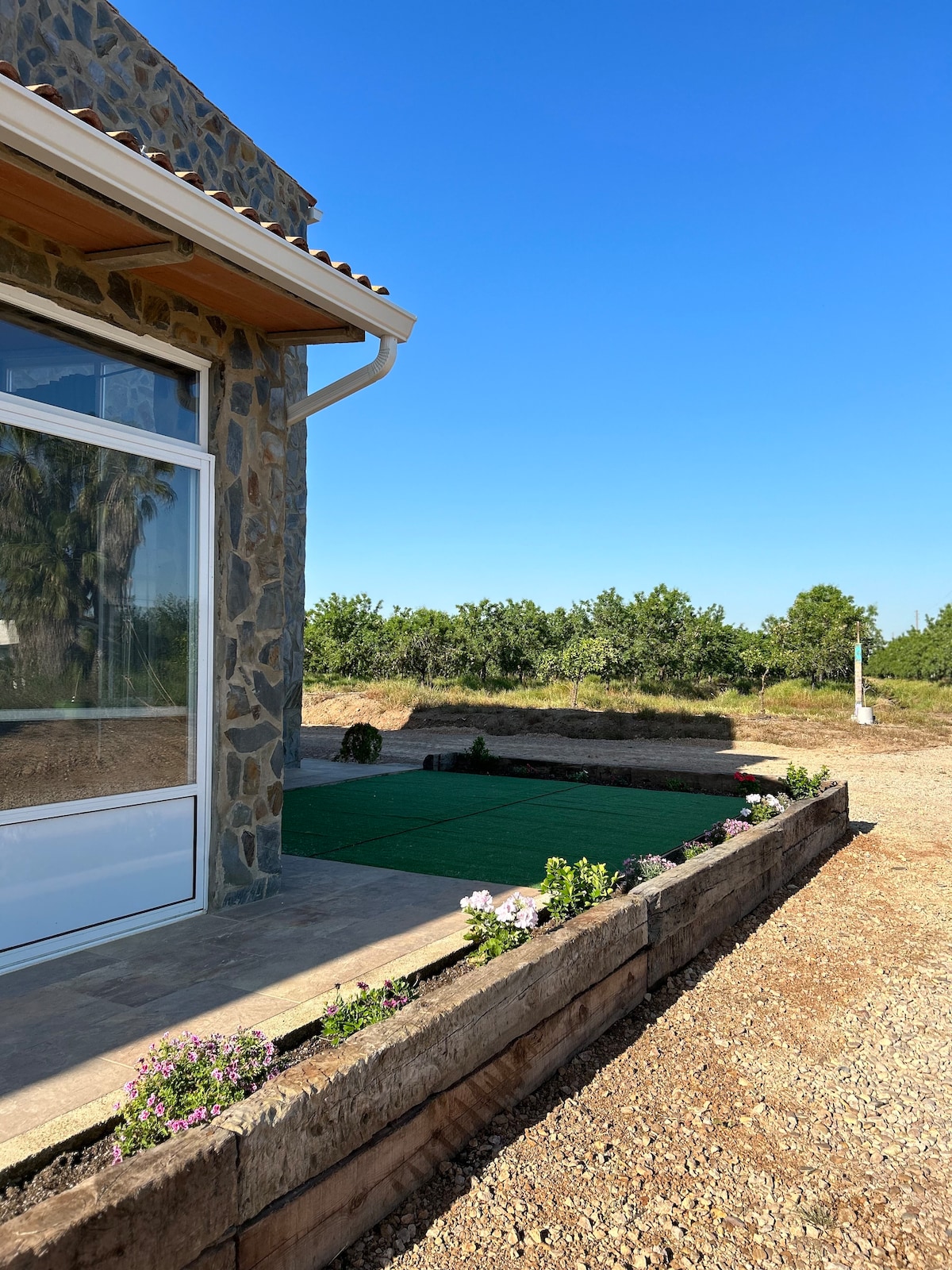 A stone exterior is featured on the building, showcasing large glass doors that open to the outdoors. A landscaped area with green grass and blooming flowers is visible, enhancing the serene surroundings.
