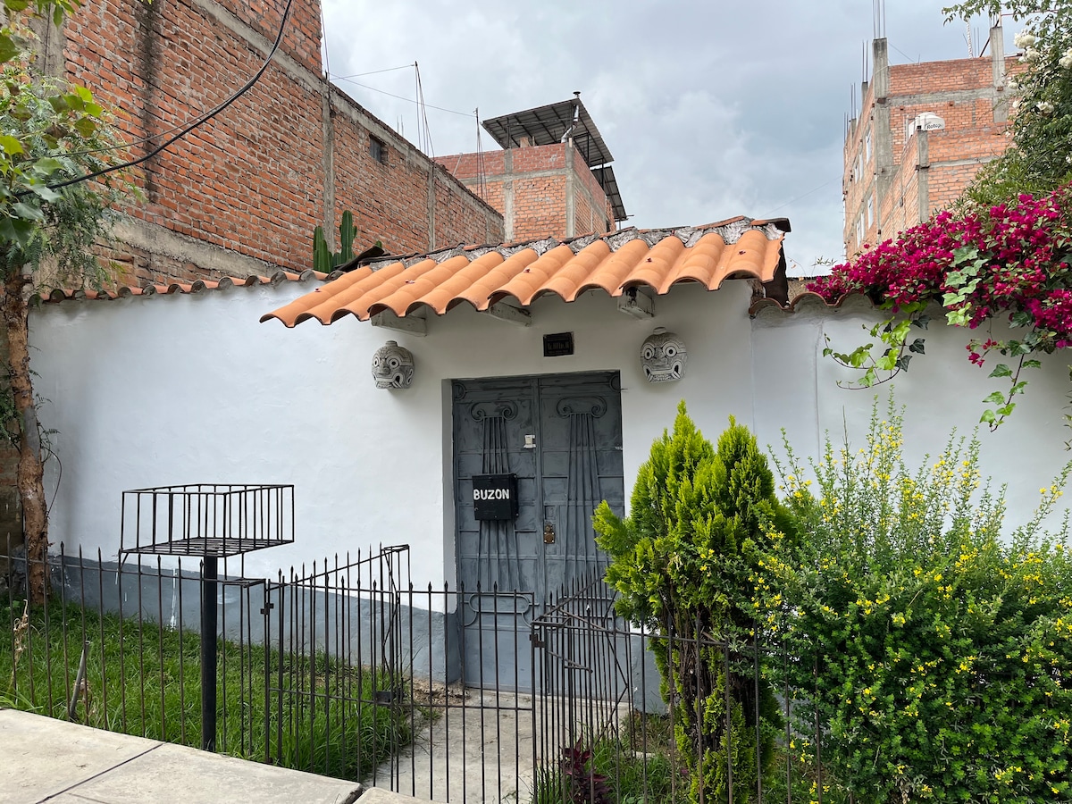 The entrance of a bungalow is framed by a low wrought iron fence and lush greenery. A wooden door with a decorative design is centered under a tiled roof, flanked by bright flowering plants. The exterior walls are painted white, enhancing the inviting appearance.