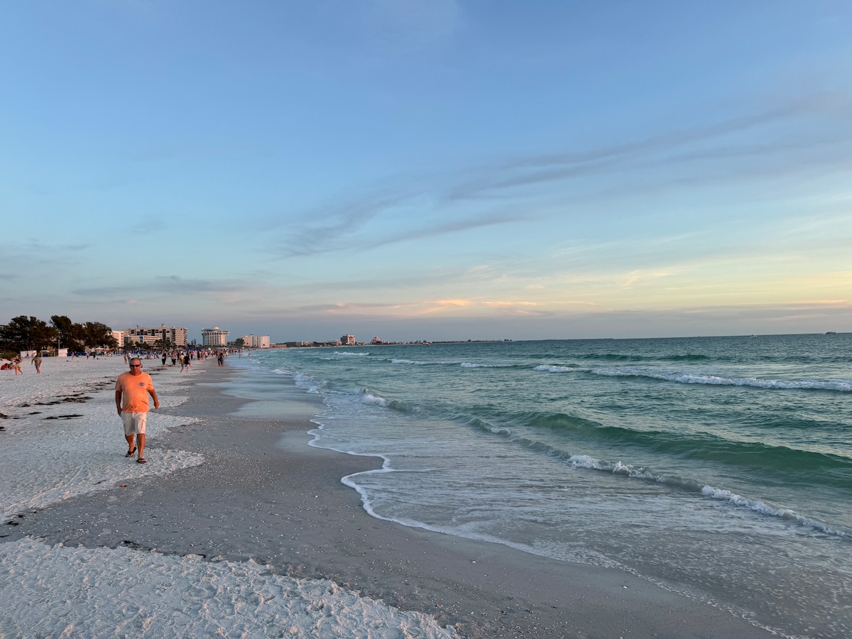 A serene beach scene features gentle waves lapping at the shore under a pastel sky. The shoreline is dotted with people enjoying the evening, while distant buildings frame the background. Soft sand stretches to the horizon, creating a tranquil atmosphere.