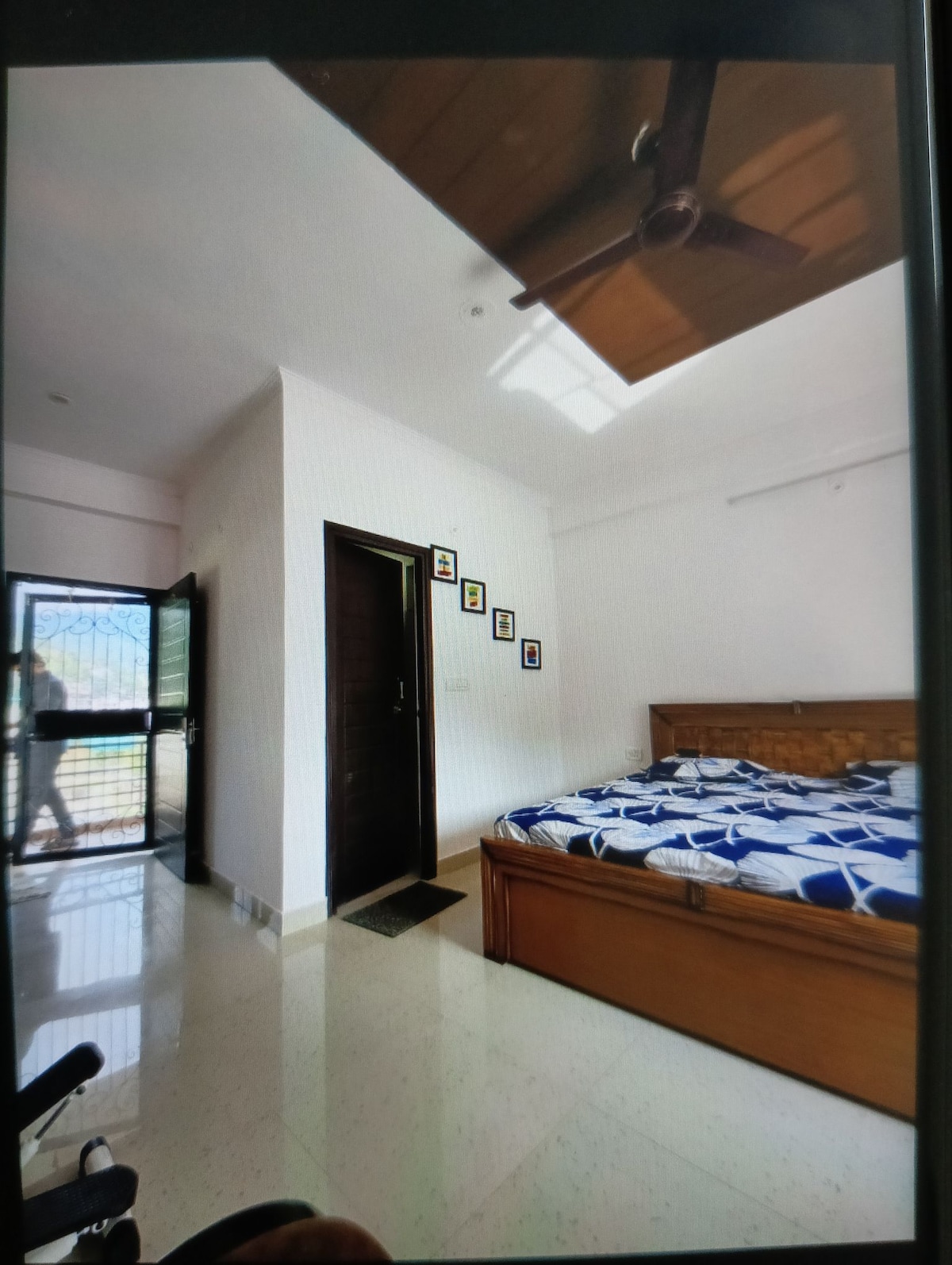 A well-lit bedroom features a spacious bed with a blue and white patterned bedspread. A dark wooden wardrobe is located near a door leading outside, while framed photographs adorn the wall. Natural light enters through a nearby window, emphasized by a ceiling fan overhead.