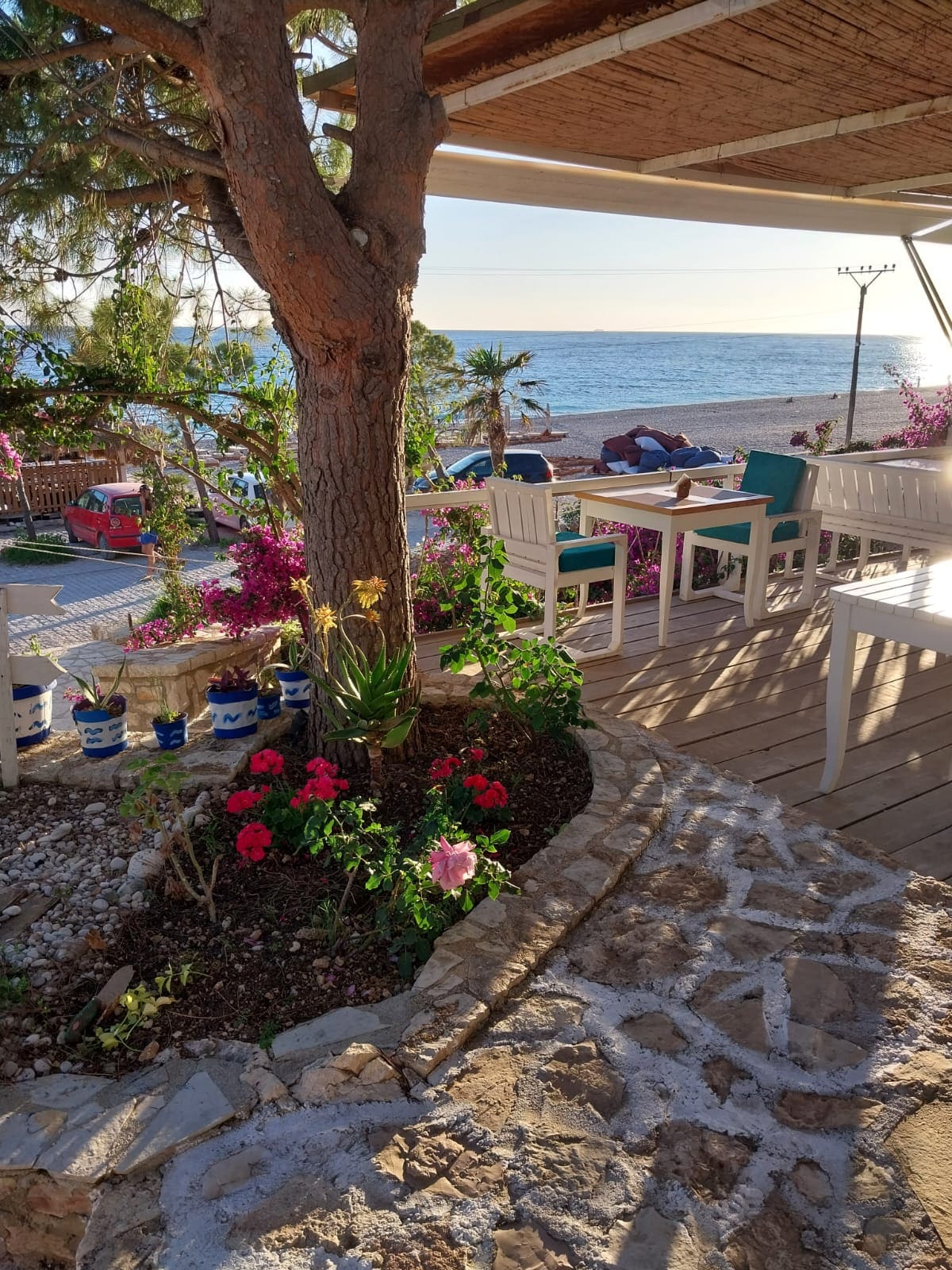 A view of a charming outdoor seating area features white chairs and a table, positioned under a pergola. Lush greenery, including a tree and colorful flowers, enhances the space. The sea sparkles in the background, providing a serene atmosphere.