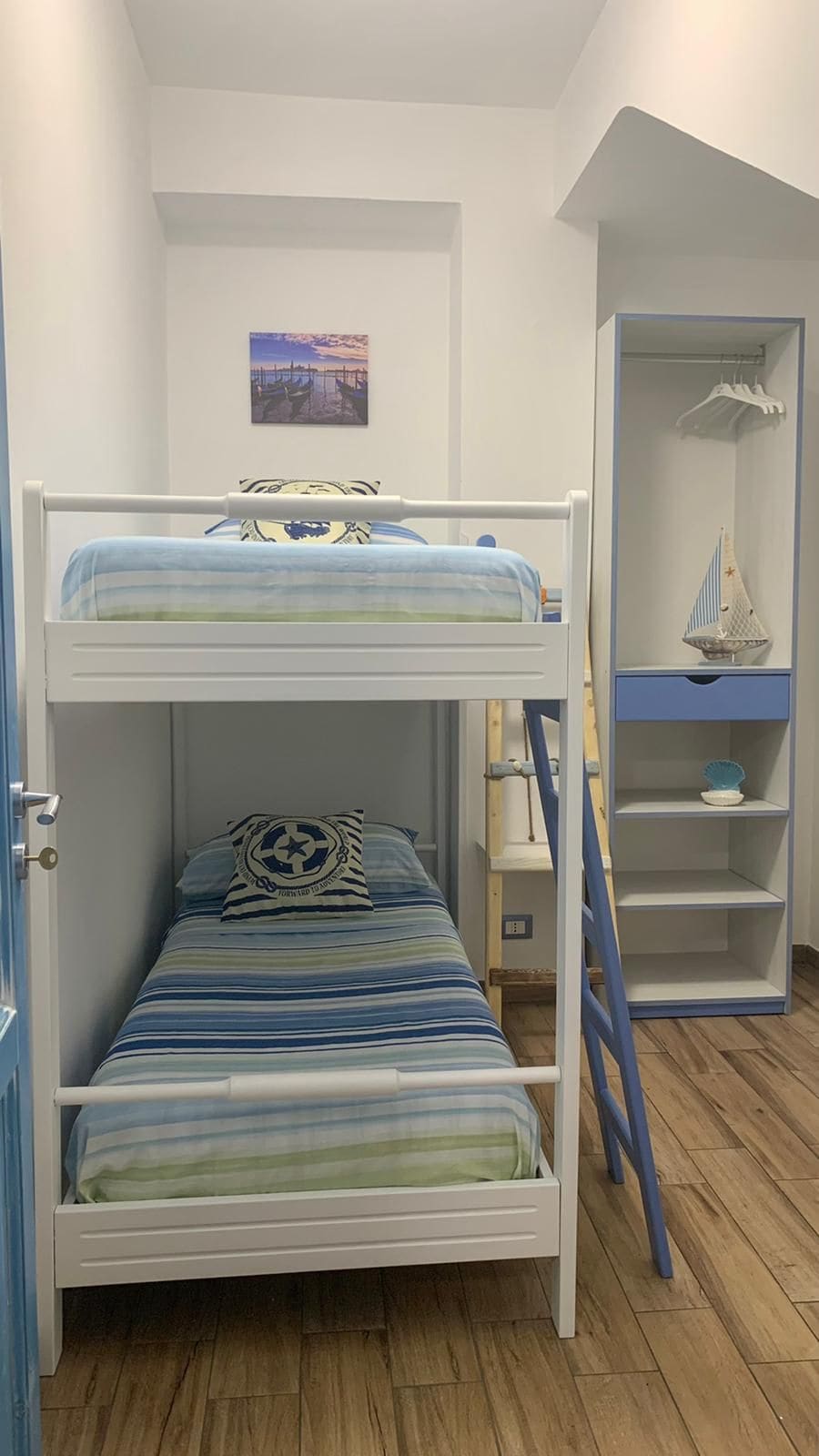 A compact bedroom featuring a bunk bed with soft blue and green striped bedding. A decorative pillow accents the upper bunk. A small shelf is mounted on the wall, housing a decorative sailboat and hanging space for clothing. Light enters through a window, enhancing the serene atmosphere.
