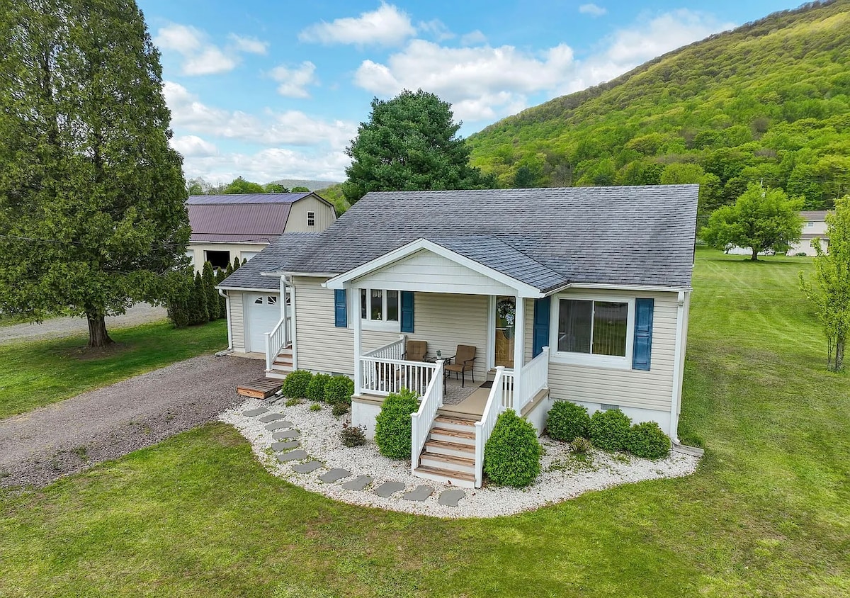 The cottage exterior is framed by lush green hills, with a welcoming front porch featuring light-colored rocking chairs. A stone pathway leads to the entrance, surrounded by neatly trimmed bushes and a manicured lawn. The two-tone siding is complemented by blue shutters.