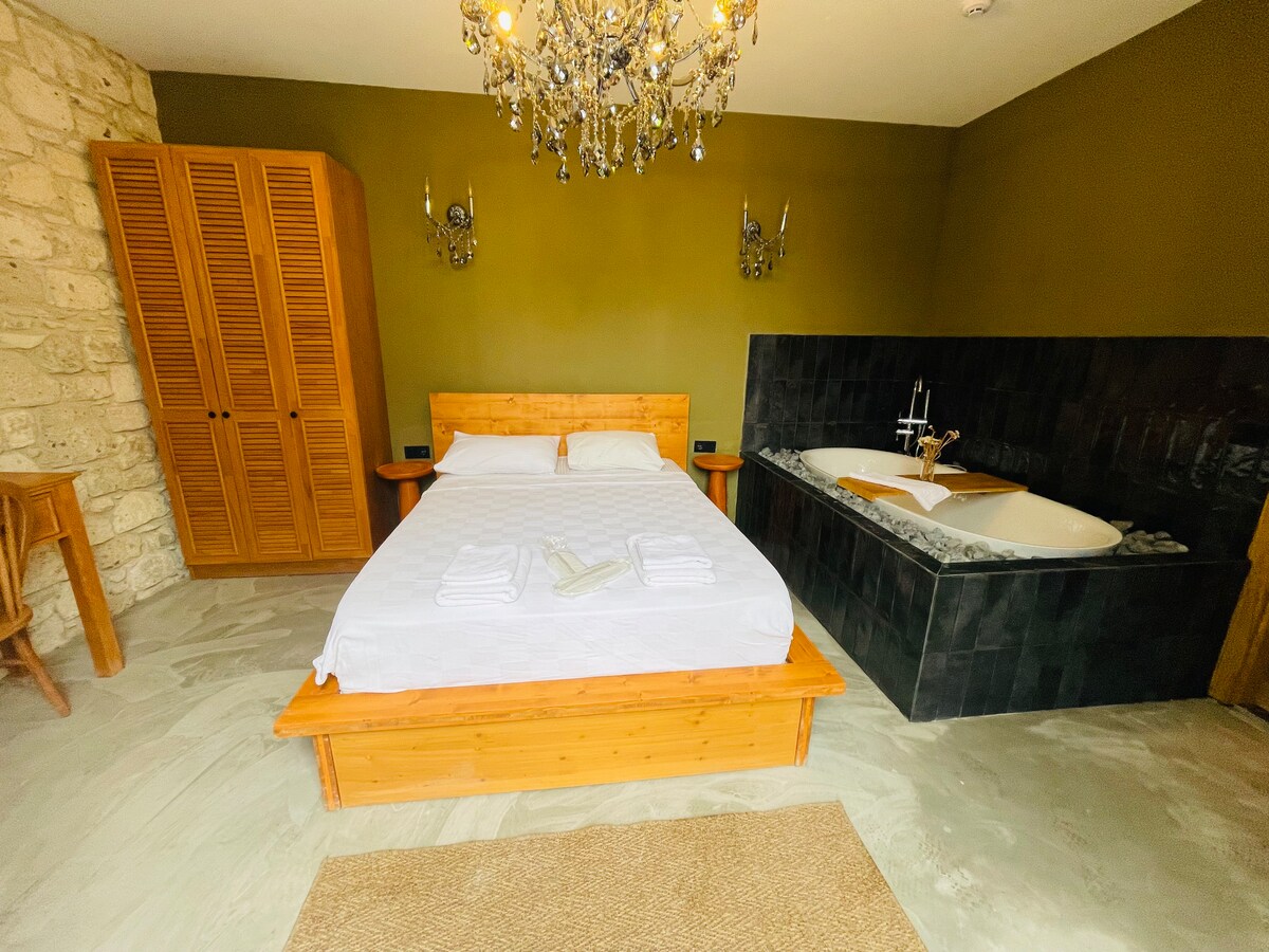 A spacious room features a wooden platform bed covered with white linens, accompanied by neatly arranged towels. Natural stone walls provide texture, while a striking chandelier hangs above. A sleek black bathtub is positioned near the bed, enhancing the room's charm. A wooden wardrobe offers ample storage.