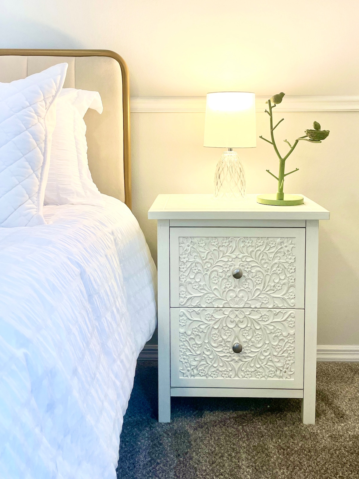 A light-colored nightstand with two intricately designed drawers stands beside a spacious bed dressed in white linens. A clear lamp with a soft glow and a small plant are positioned on top of the nightstand, adding subtle details to the serene ambiance.