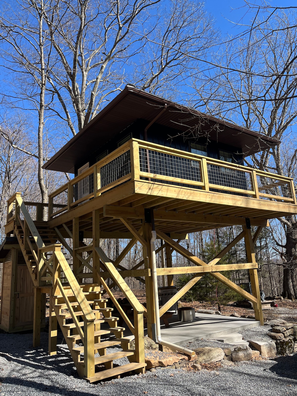 A wooden cabin elevated on a sturdy platform is surrounded by trees, showcasing a sloped roof and spacious balcony. Stairs lead up to the entrance, providing access to the airy outdoor area. A stone fire pit is located beneath the cabin, enhancing the natural setting.