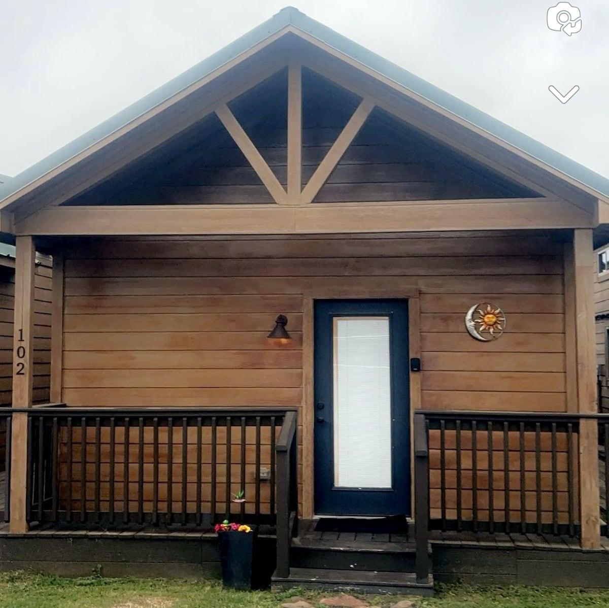 A quaint cabin exterior showcases natural wooden siding and a gabled roof. A welcoming front porch is adorned with a small flower planter, while a decorative wall piece adds a touch of charm beside the entrance door.