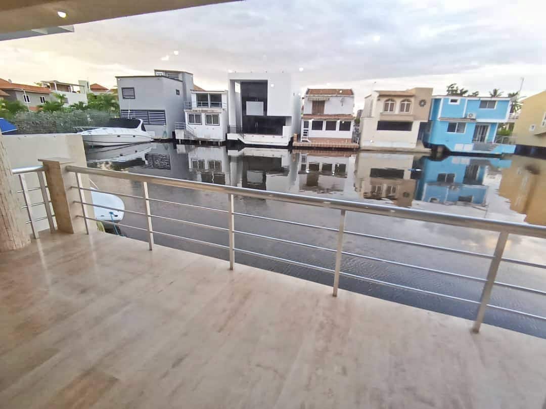 A spacious balcony overlooks a calm waterway, where colorful homes line the opposite shore. The railing is sleek and modern, providing an unobstructed view of the mirrored reflections on the water's surface, enhancing the serene environment.