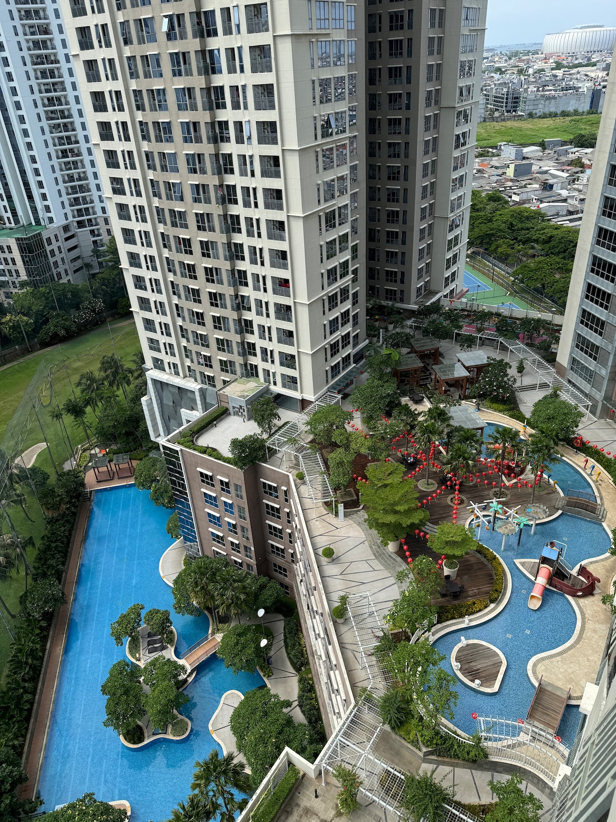 An aerial view showcases a well-maintained outdoor space featuring a swimming pool and a playground surrounded by lush greenery. Vistas of the surrounding buildings and cityscape are visible in the background, highlighting the apartment's central location.