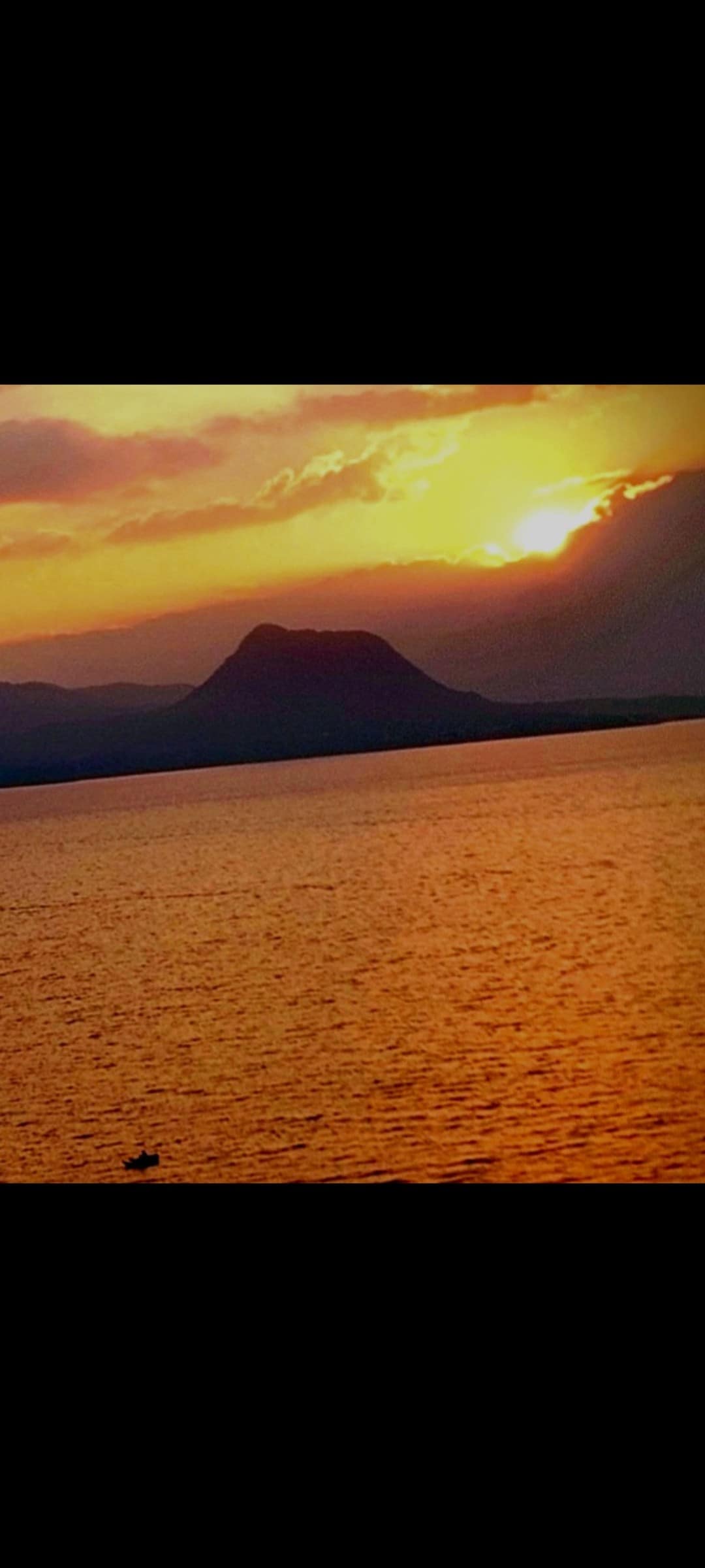 A serene sunset is captured over the lake, casting warm hues of orange and yellow across the water. Silhouetted mountains outline the horizon, creating a tranquil scene of natural beauty.