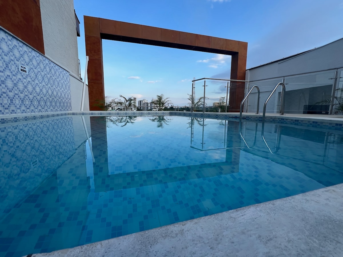 A tranquil rooftop swimming pool is framed by a modern architectural structure. The clear blue water reflects the sky, offering a serene environment. Overhead, the design creates an open area for panoramic city views and peaceful relaxation.