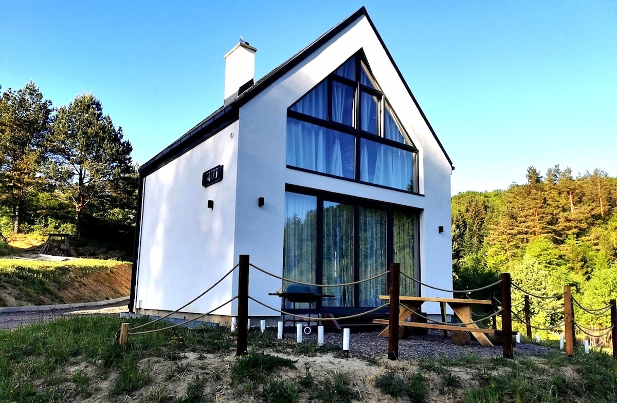 A modern two-story house is set against a backdrop of trees. The structure features large glass windows that allow natural light to fill the space. A railing made of wooden posts and rope outlines the entrance, enhancing the connection to the surrounding landscape.