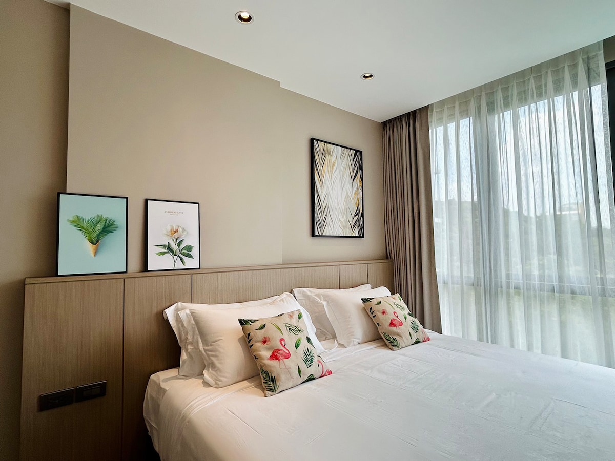 A serene sleeping area is featured, with a spacious bed adorned with decorative pillows. Light-colored artwork hangs on the wall beside the bed, and large windows allow natural light to fill the room, complemented by soft, sheer curtains.