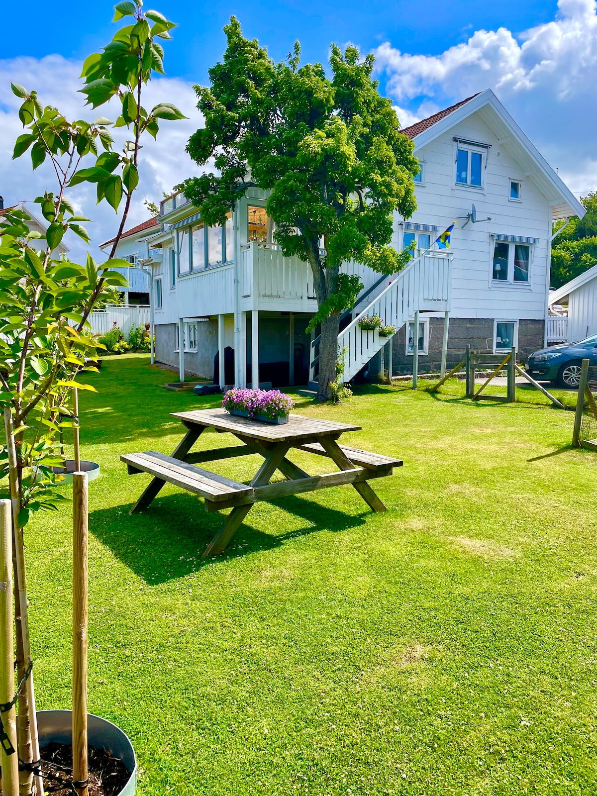 A white two-story house is situated in a well-maintained yard, featuring a neatly mowed lawn. A wooden picnic table with a flower arrangement is positioned beneath a tree, inviting outdoor gatherings. The house is surrounded by simple landscaping, creating an open and welcoming environment.