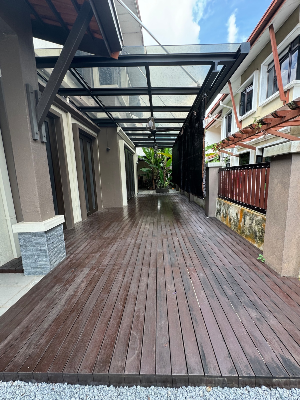 An outdoor deck features polished wooden flooring, extending toward a garden area with lush greenery. A transparent roof structure above allows natural light to fill the space while providing shelter. The clean lines and spacious layout create an inviting area for relaxation and gatherings.