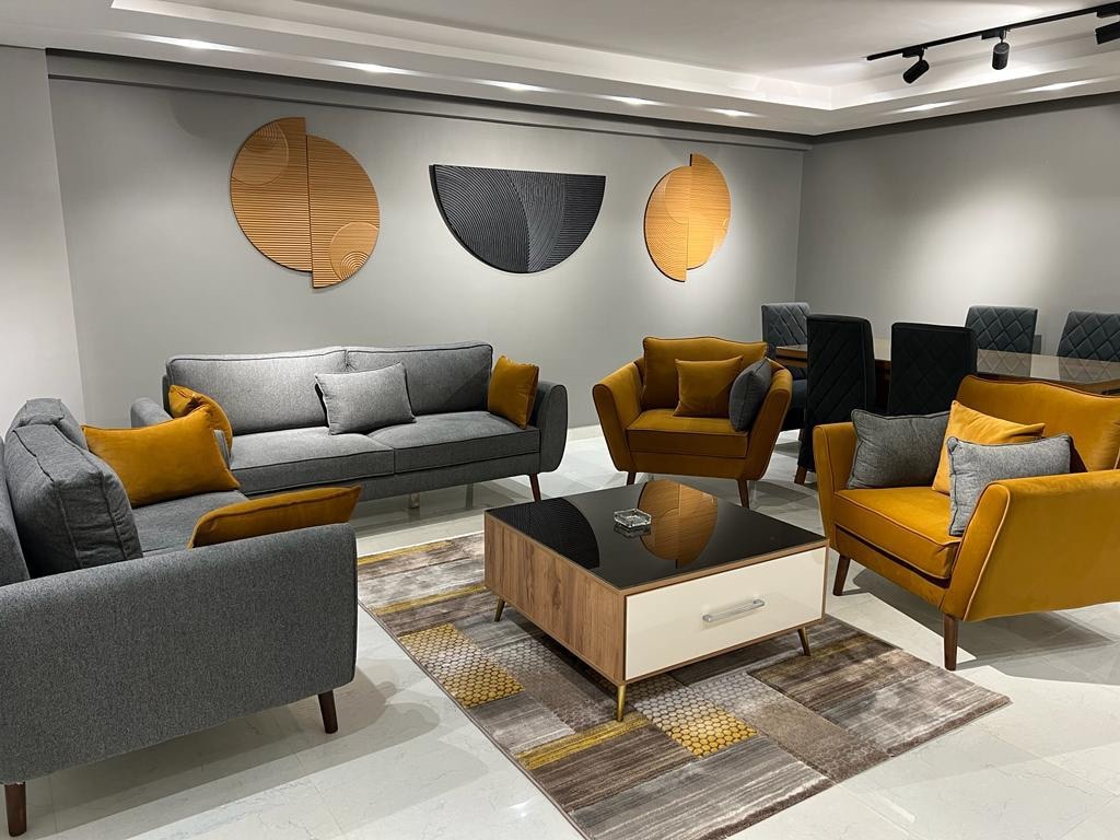 The living area features a modern arrangement of gray and mustard-colored furniture. Two stylish armchairs and a sofa are complemented by a central coffee table, resting on a patterned area rug. Wall art composed of wooden shapes adds a contemporary touch to the space.