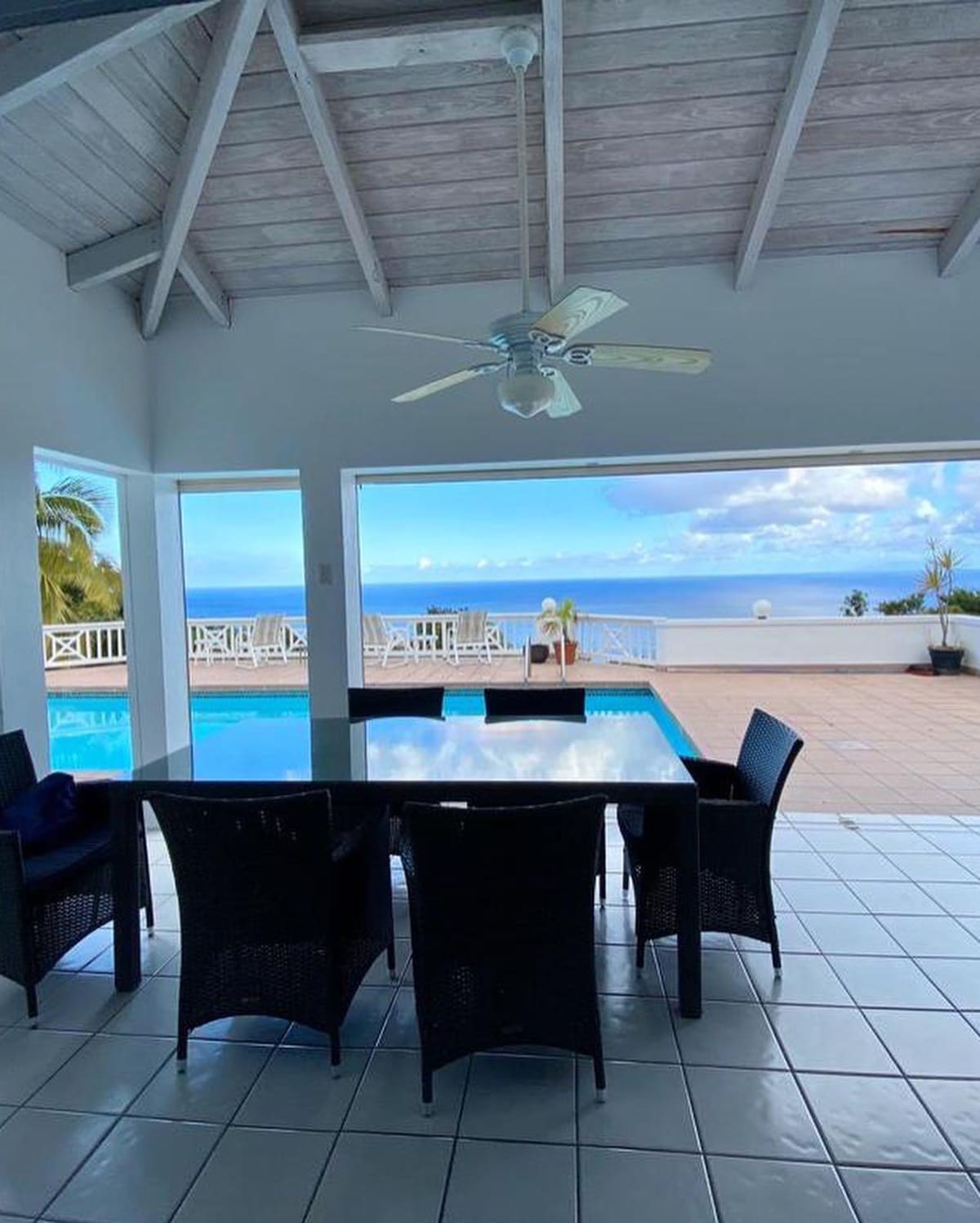 A dining space is presented with dark wicker furniture, set beneath a ceiling fan and framed by large glass doors. The view reveals an outdoor pool and the Caribbean Sea beyond, complemented by bright blue skies and distant clouds.