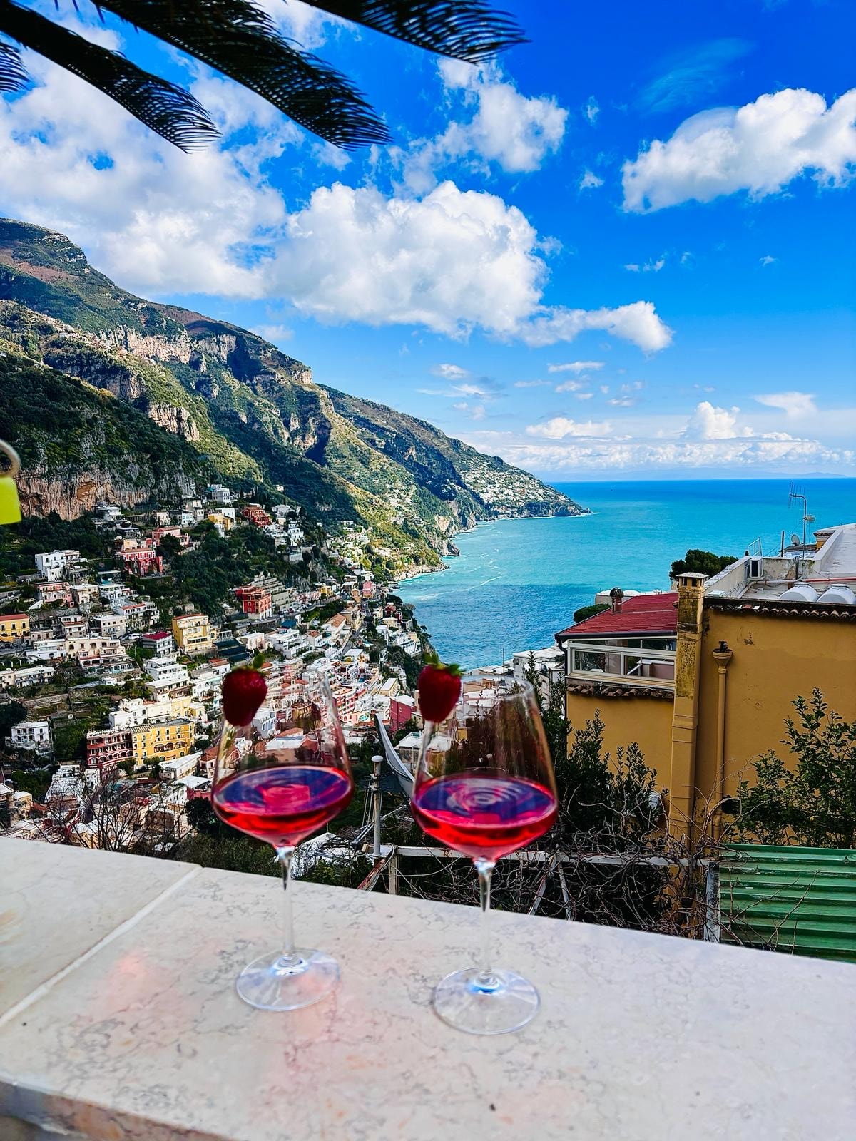 Two glasses filled with red drinks are positioned on a countertop. In the background, a stunning view of Positano's colorful hillside houses and the clear blue sea is visible, framed by lush green mountains and a bright blue sky with scattered clouds.