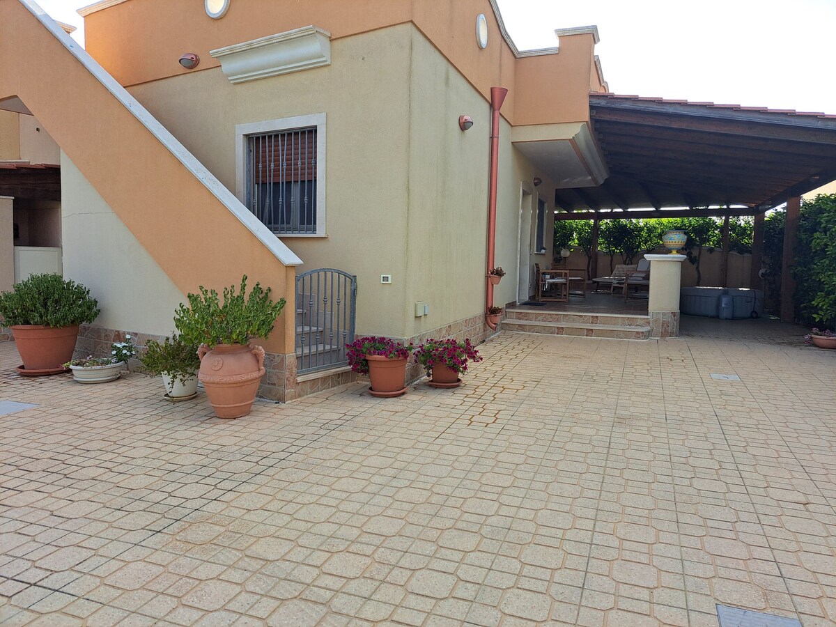 The exterior of the villa features a spacious tiled patio, bordered by an array of potted plants. A staircase leads to the upper level, while a shaded seating area is visible under a wooden roof, creating a welcoming space for relaxation.