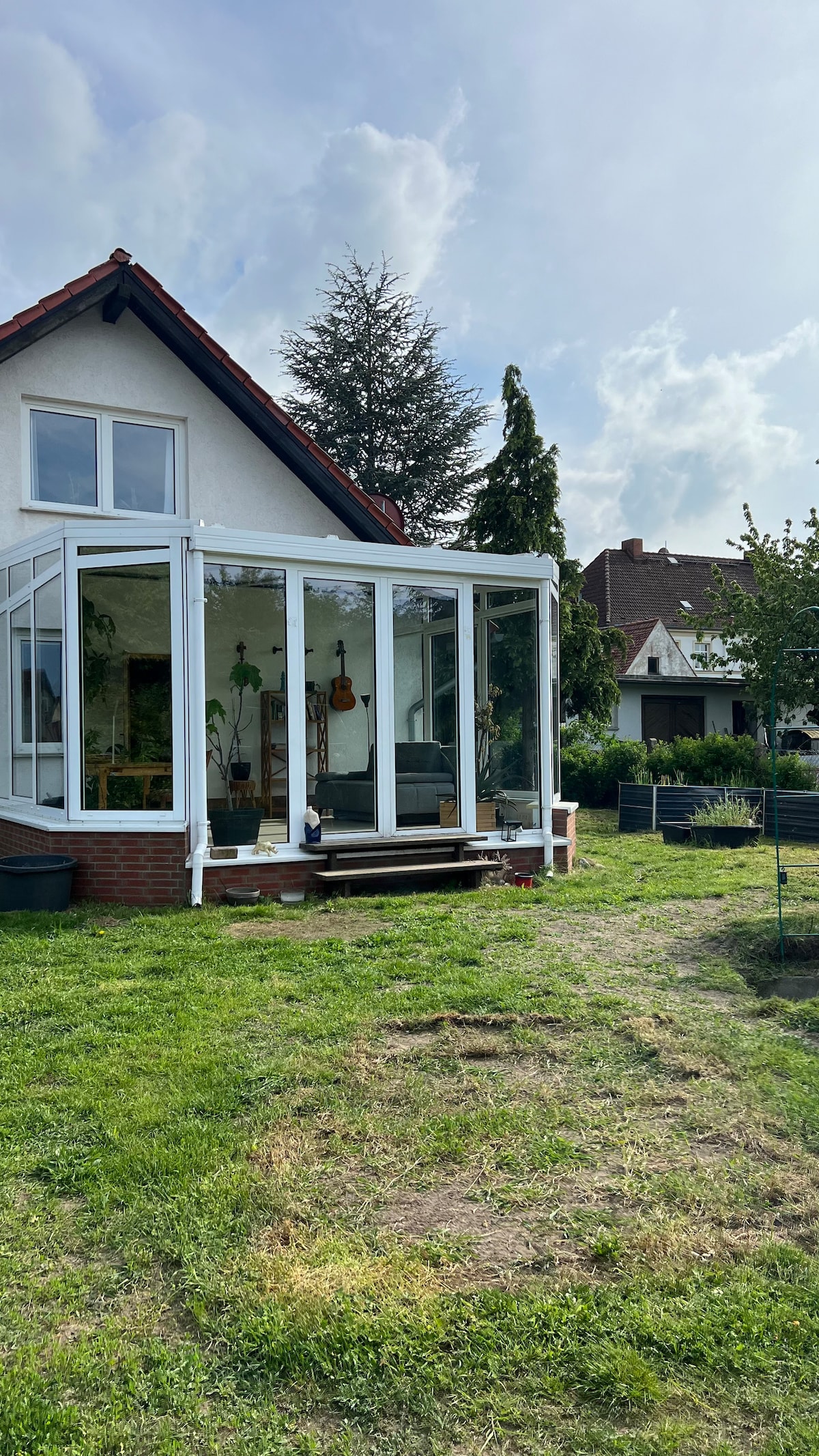A sunlit conservatory is attached to the house, featuring large glass panels that invite natural light. The surrounding garden is lush and green, with a spacious lawn and trees providing shade in the background. The exterior reflects a blend of modern and traditional architecture.