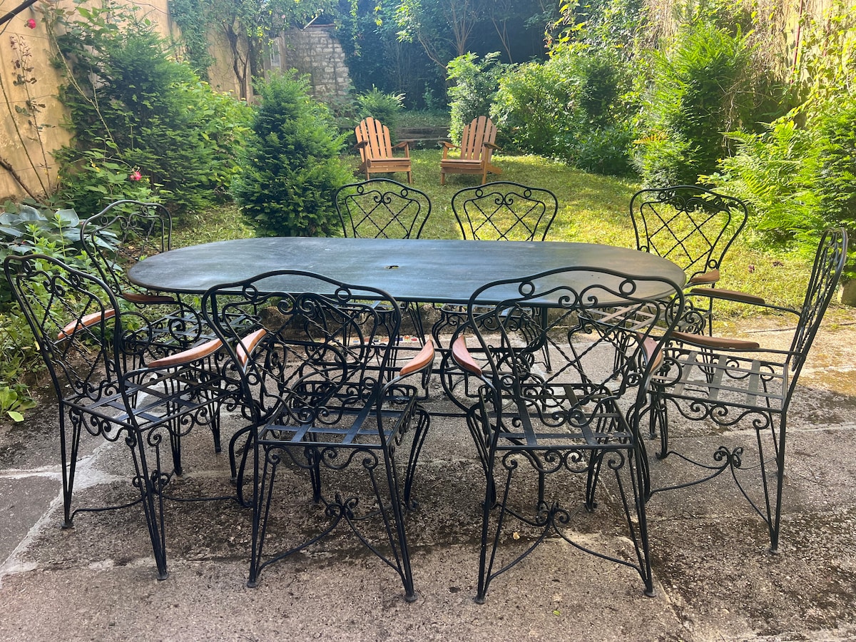 A round dining table is surrounded by multiple wrought-iron chairs in a tranquil garden setting. Lush greenery and tall hedges create a private atmosphere, while a stone path leads through the inviting outdoor space, perfect for gatherings and relaxation.