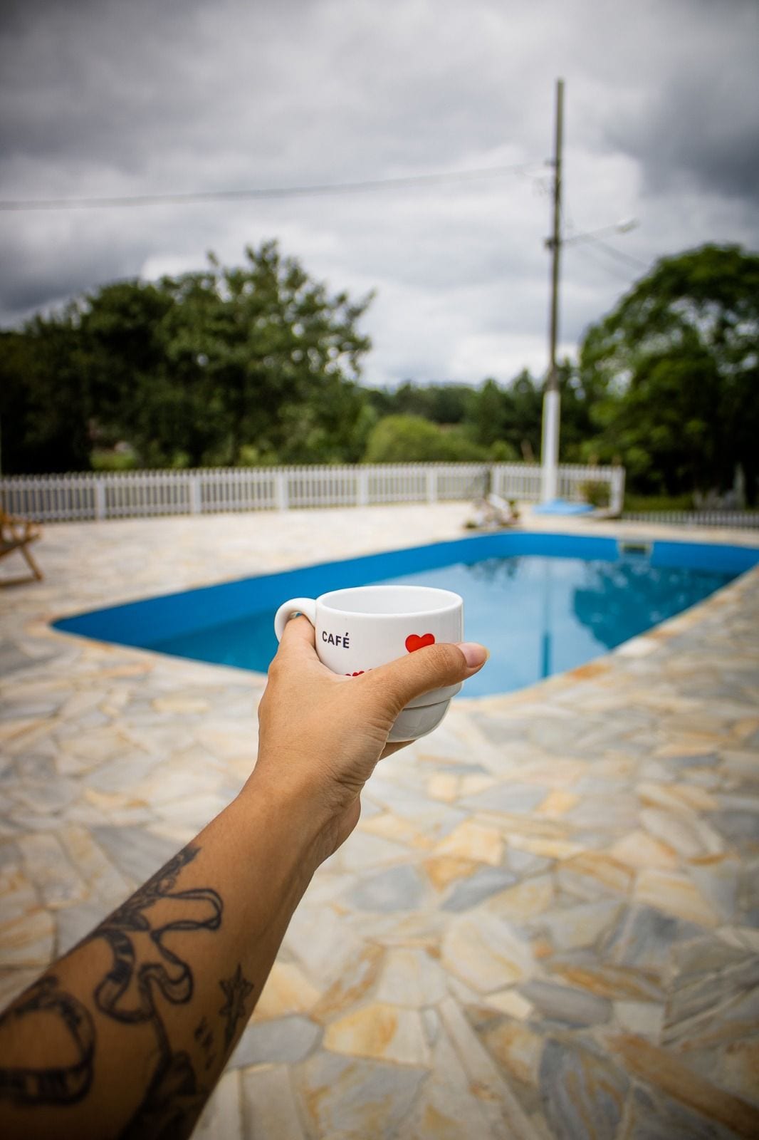 A person holds a coffee cup in front of a serene pool area. The pool's turquoise water contrasts with the light stone flooring. Lush trees and greenery frame the space, creating a natural setting. A white railing surrounds the pool, enhancing the outdoor ambiance.