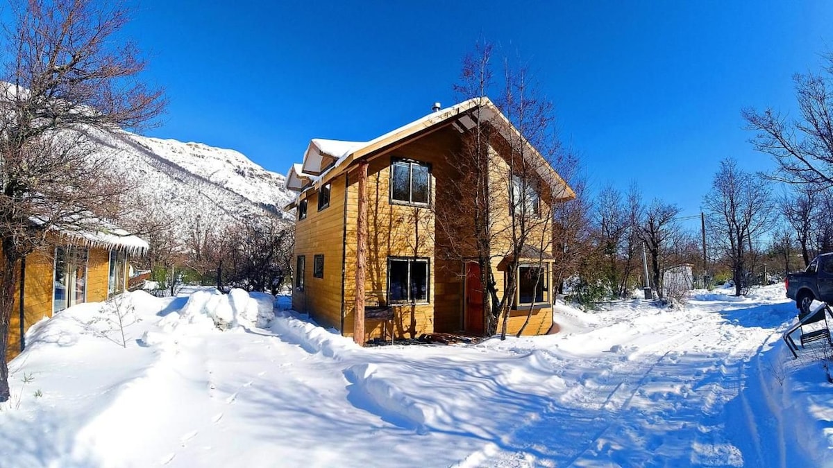 A two-story wooden building is set amidst a snowy landscape, surrounded by trees. Sunlight reflects off the snow, highlighting the building's large windows. The exterior features a combination of wood and stone accents, contributing to a harmonious look with the natural surroundings.