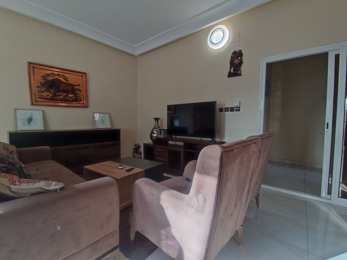 A comfortable living area features two plush armchairs and a wooden coffee table. A television is positioned on a dark wooden console, with framed artwork displayed on the wall. Sunlight streams in through a nearby window, illuminating the space.