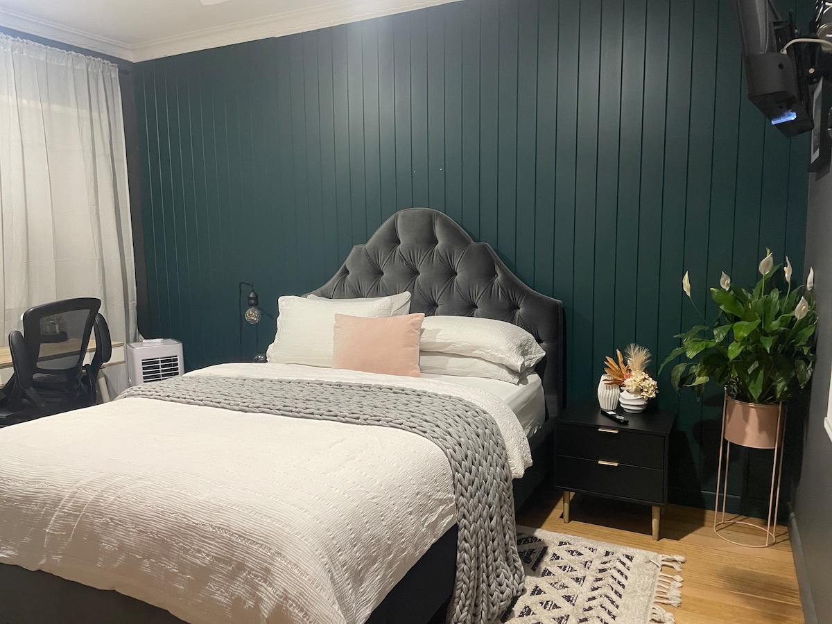 A cozy sleeping area features a large, tufted black headboard behind a spacious bed layered with white linens. A decorative throw rests across the bed, while a small side table holds potted plants. A chair and air conditioning unit complete the room’s functional decor.