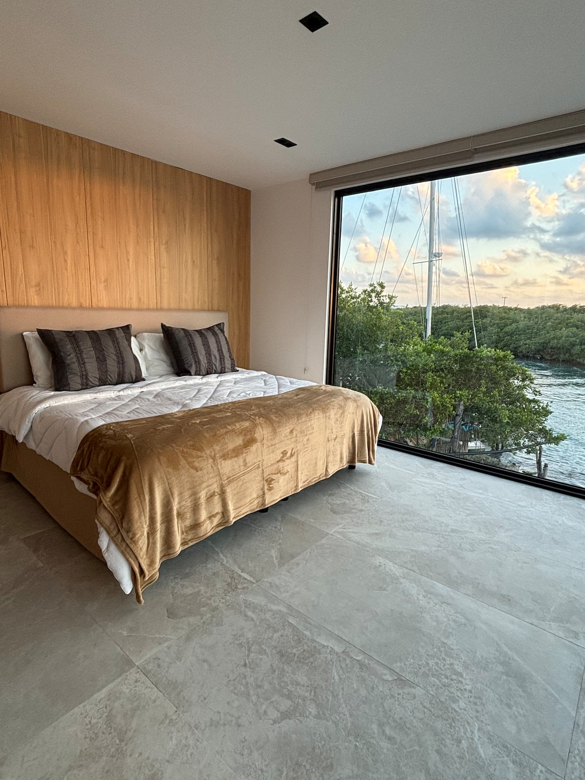A spacious sleeping area features a large bed with neutral-toned linens and decorative pillows. Floor-to-ceiling windows reveal serene views of the water and surrounding greenery, allowing natural light to illuminate the room's clean lines and contemporary design.