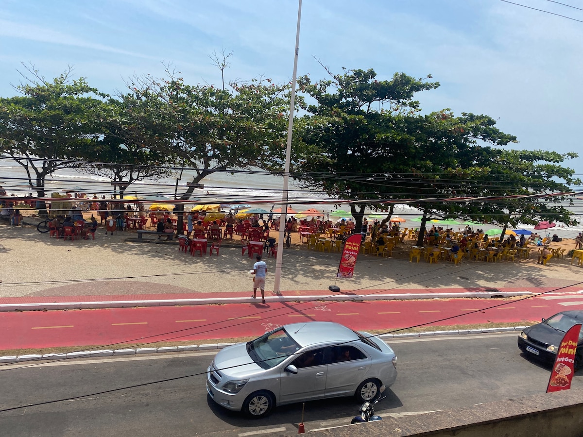 A lively beach promenade is visible, lined with vibrant yellow umbrellas and chairs under leafy trees. The sandy beach is in the distance, with people enjoying the sun. A red bike lane runs adjacent to the road, where a silver car is parked.