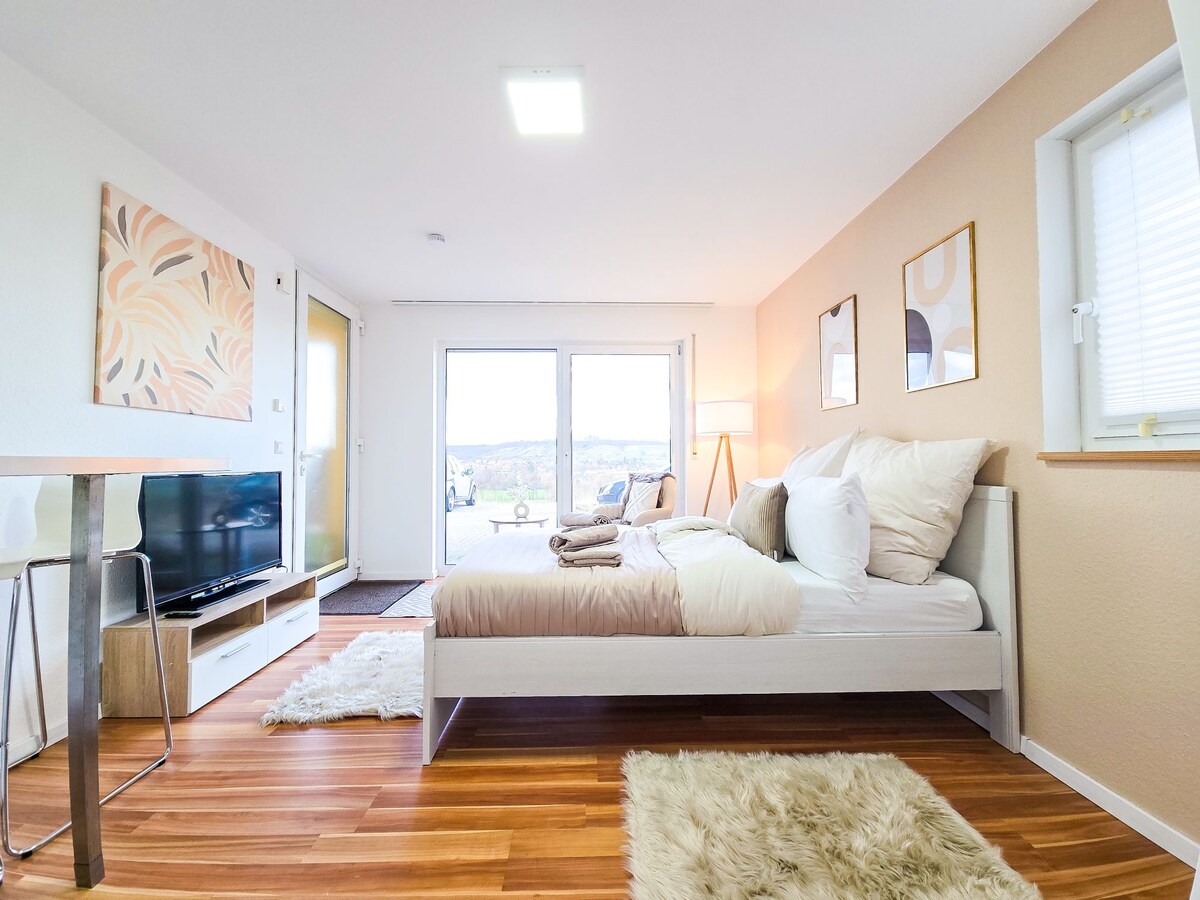 A spacious room is featured, showcasing a large bed with neutral bedding. A flat-screen TV is mounted on a wooden stand, and a small table with chairs is positioned nearby. Natural light fills the area from large glass doors leading to a balcony with scenic views.