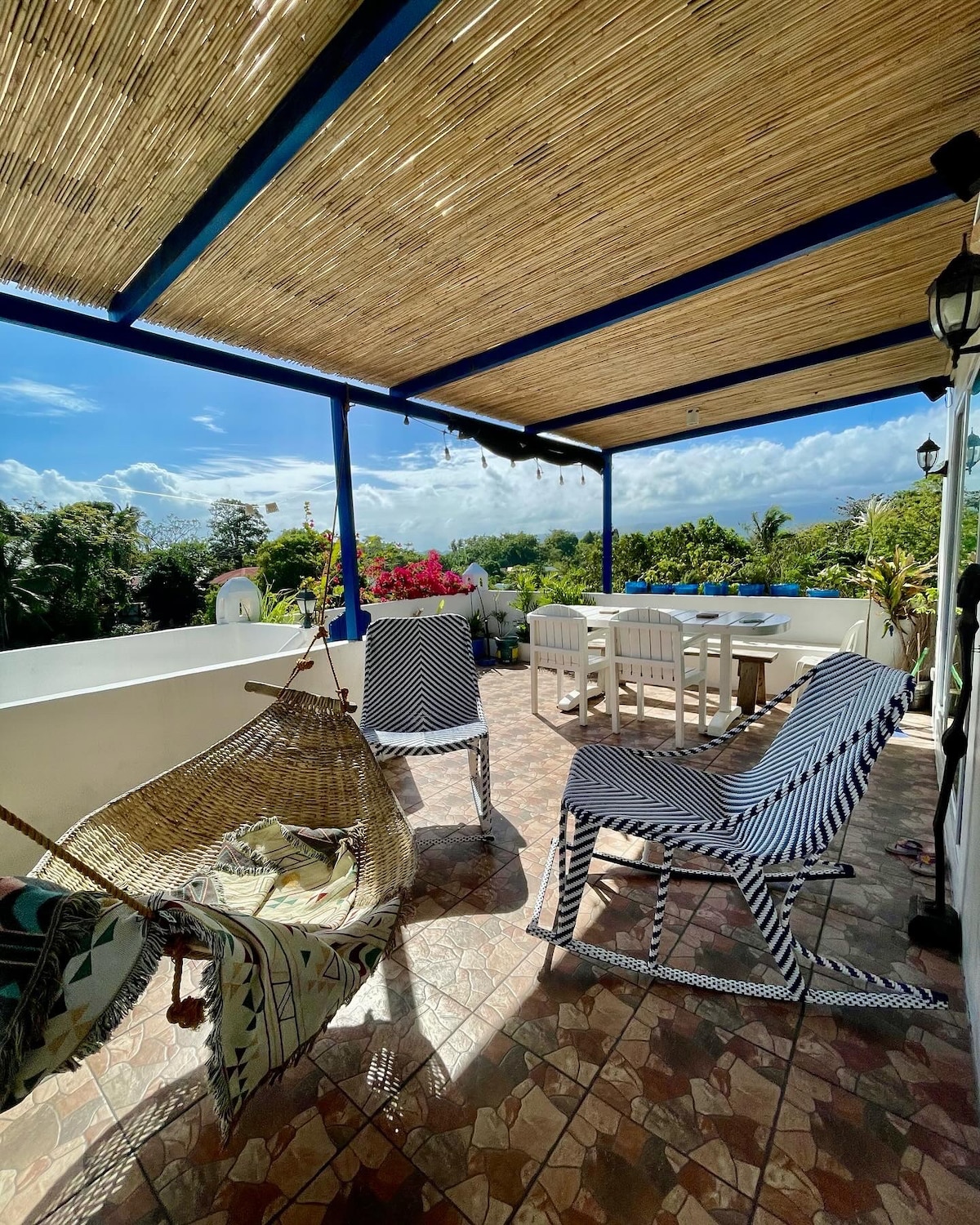 An outdoor terrace is presented, featuring stylish striped seating and a wooden dining table. Potted plants add greenery to the space, and a hammock is visible for relaxation. A scenic view of lush vegetation and distant hills is framed by a sunshade overhead.