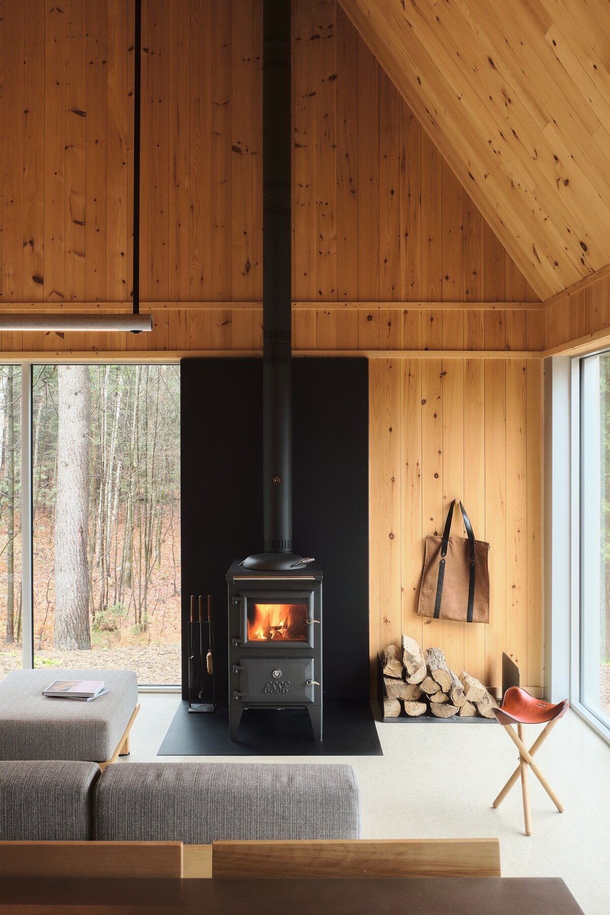 A warm living area features a wood-burning stove that emits a gentle glow. Natural wood paneling complements large windows that offer views of the surrounding forest. A cozy gray sofa and a wooden chair are arranged nearby, inviting relaxation in a minimalist setting.