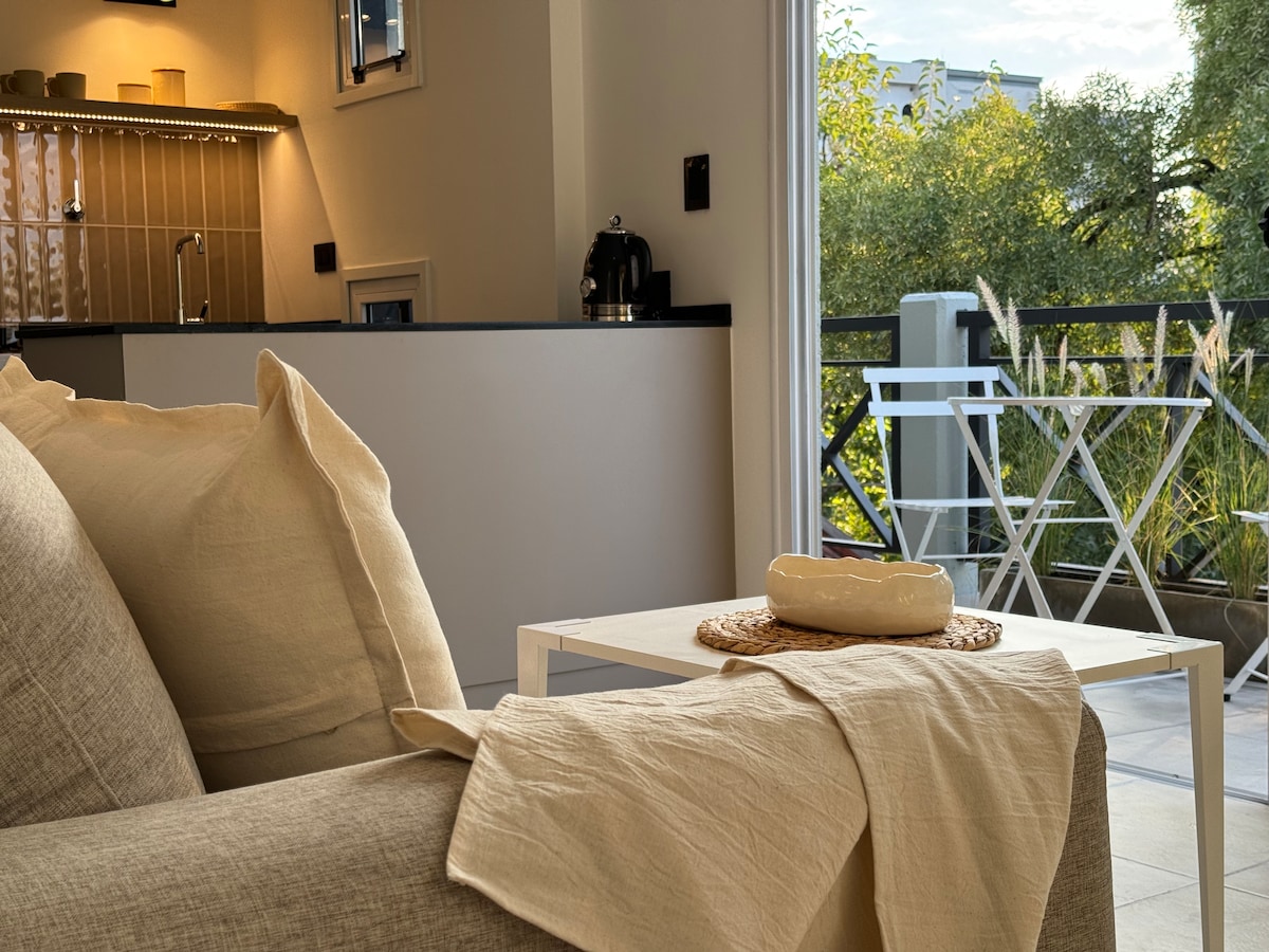 A cozy living area features a beige sofa adorned with a softly draped fabric. A minimalist table sits in front, topped with a decorative bowl. In the background, the kitchen area is visible, along with a small balcony showcasing outdoor seating and greenery.
