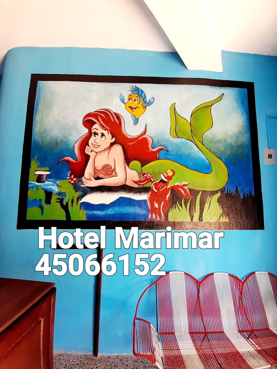 A vibrant mural adorns a blue wall, featuring a whimsical underwater scene with a mermaid and a colorful fish. A wooden dresser and a patterned seating area can be seen in the foreground, adding character to the lively space.