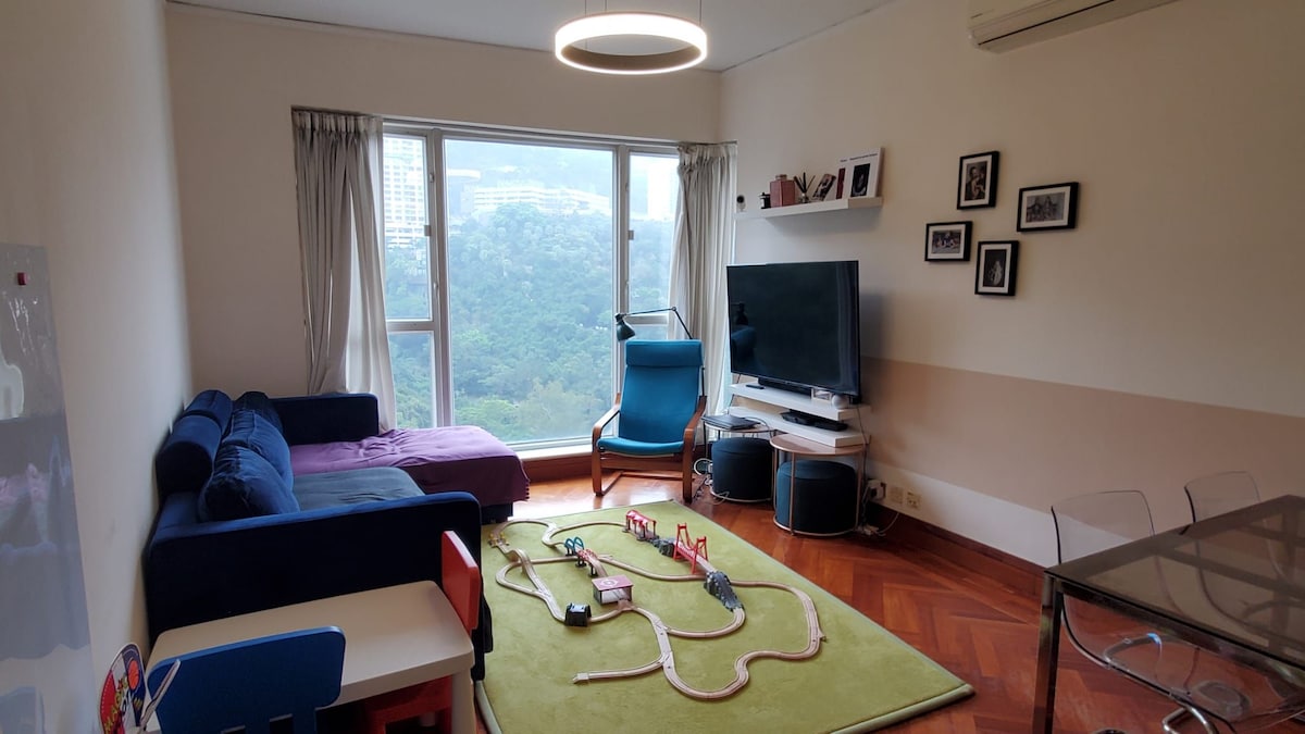 A bright living room showcases a spacious sofa in dark blue and a purple chaise lounge. A television is mounted on the wall across from a glass dining table. A green rug features a toy train set. Large windows provide an expansive view of the surrounding landscape.