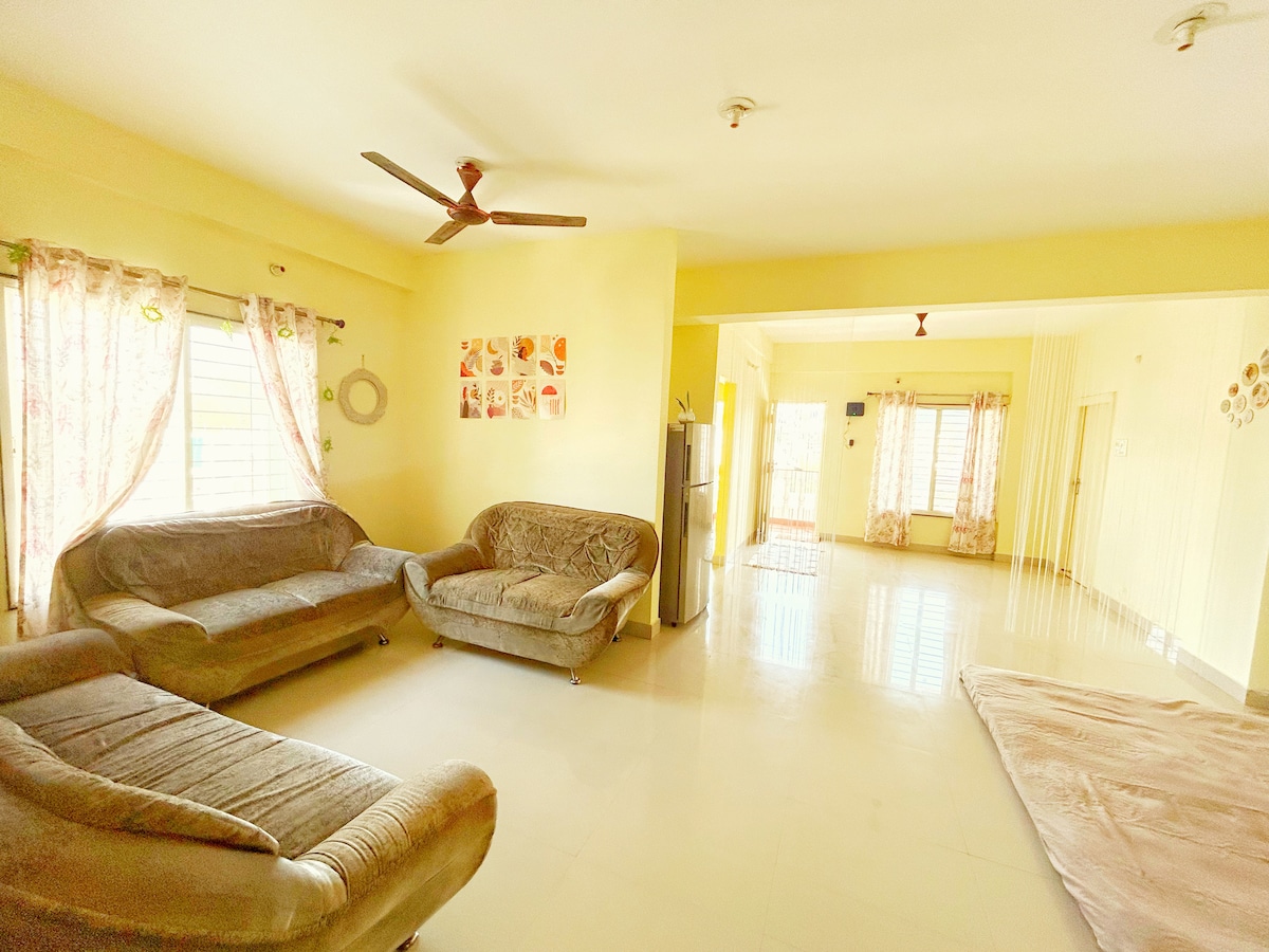 A spacious living area is displayed, featuring light yellow walls and ample natural light. Two comfortable sofas are positioned around a soft area rug, and a ceiling fan is visible above. Decorative curtains frame the windows, contributing to the airy atmosphere.