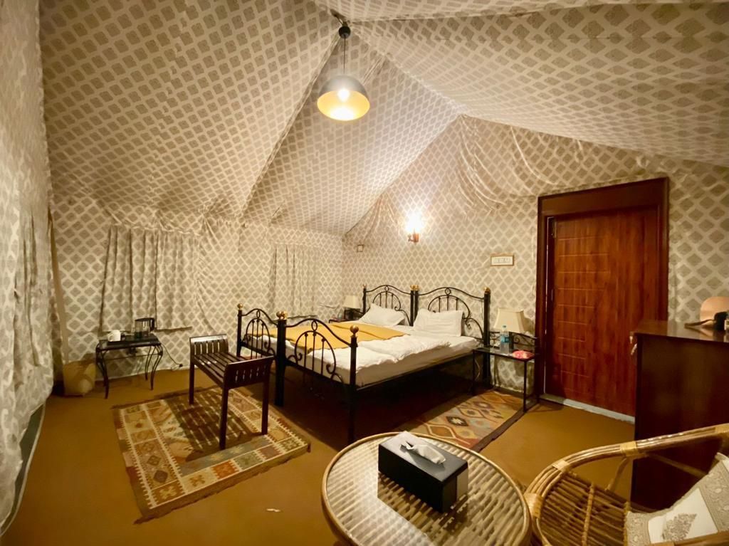 The room features a spacious bed surrounded by decorative fabric drapes, creating a unique ambiance. A small wooden table and chairs are positioned beside the bed, while a cabinet and plush rugs add to the room's comfort and style.